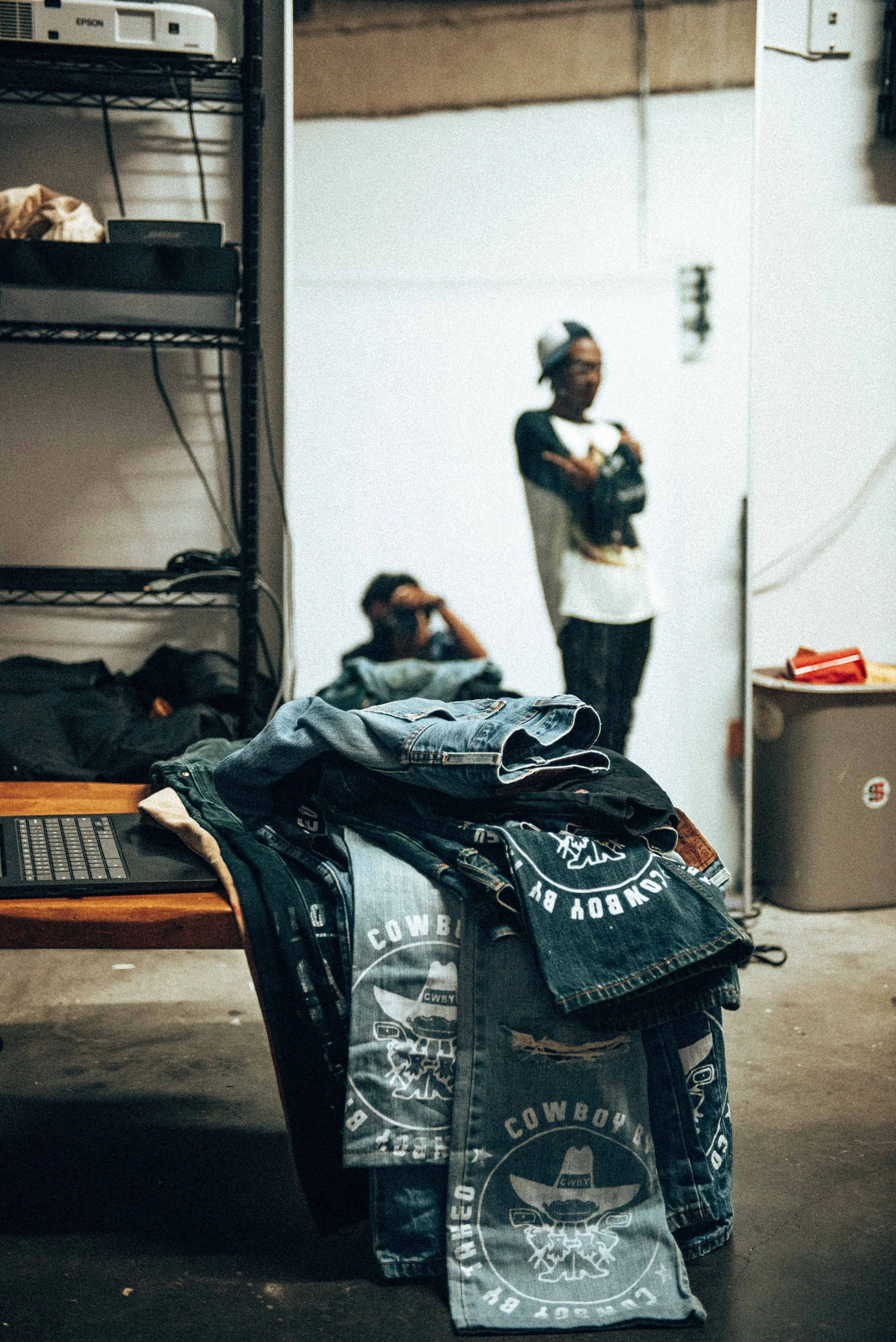 Clothes, including denim jackets with cowboy logos, are draped over a table in a room with metal shelving and a laptop. In the background, two people are visible, one seated and covering their face, the other standing with crossed arms and wearing a cap and glasses.