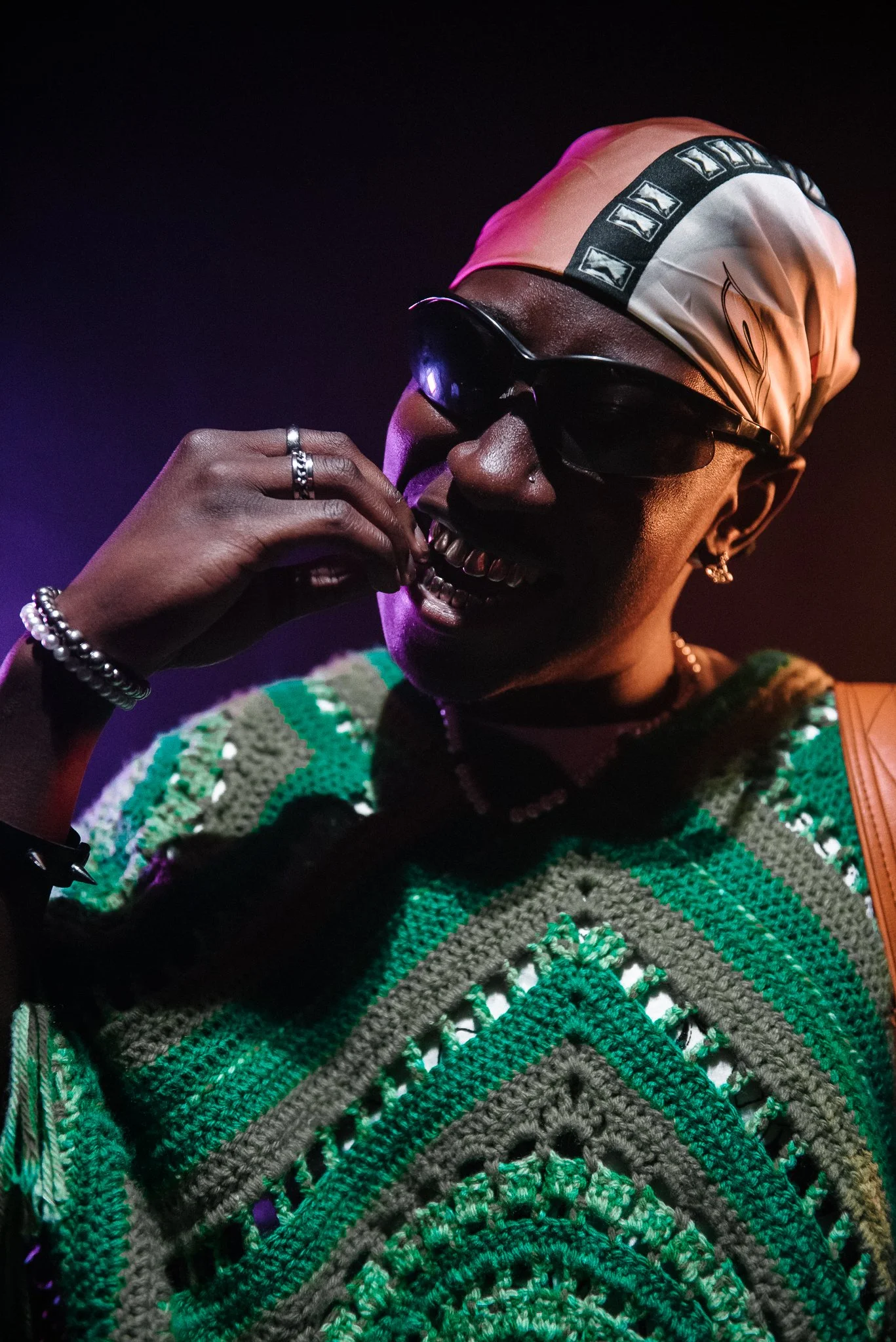 Person wearing sunglasses, a colorful crochet top, and a patterned headscarf, smiling and showing their teeth.
