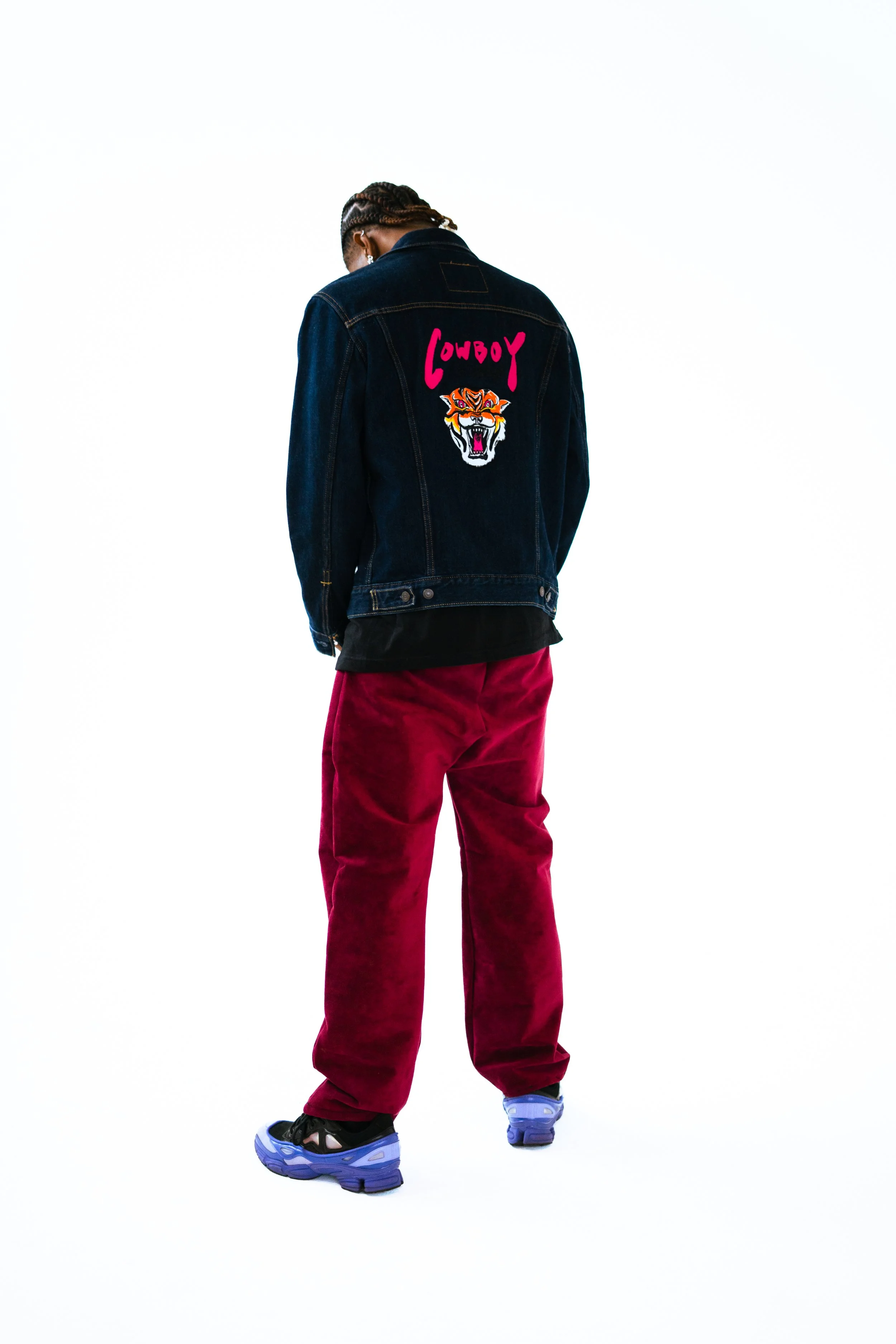 Person wearing a dark denim jacket with a tiger embroidery and the word 'Cowboy' on the back, red velvet pants, black and purple sneakers, standing against a white background.