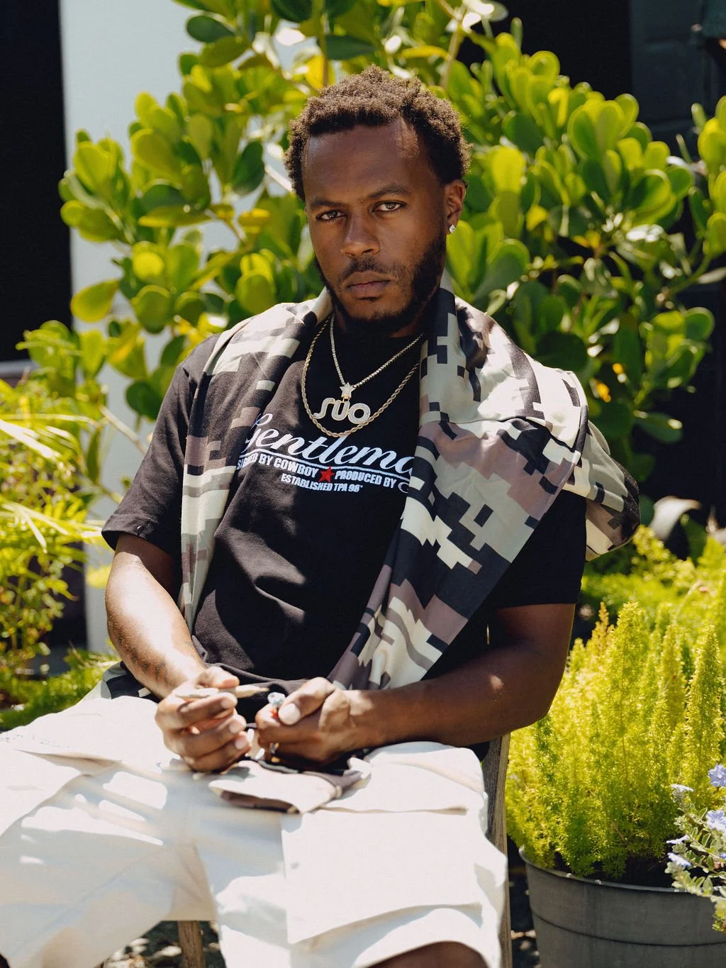A man sitting outdoors at a table with lush green plants in the background, wearing a black T-shirt with white lettering, layered with a camouflage-patterned shirt, and accessorized with necklaces and earrings.