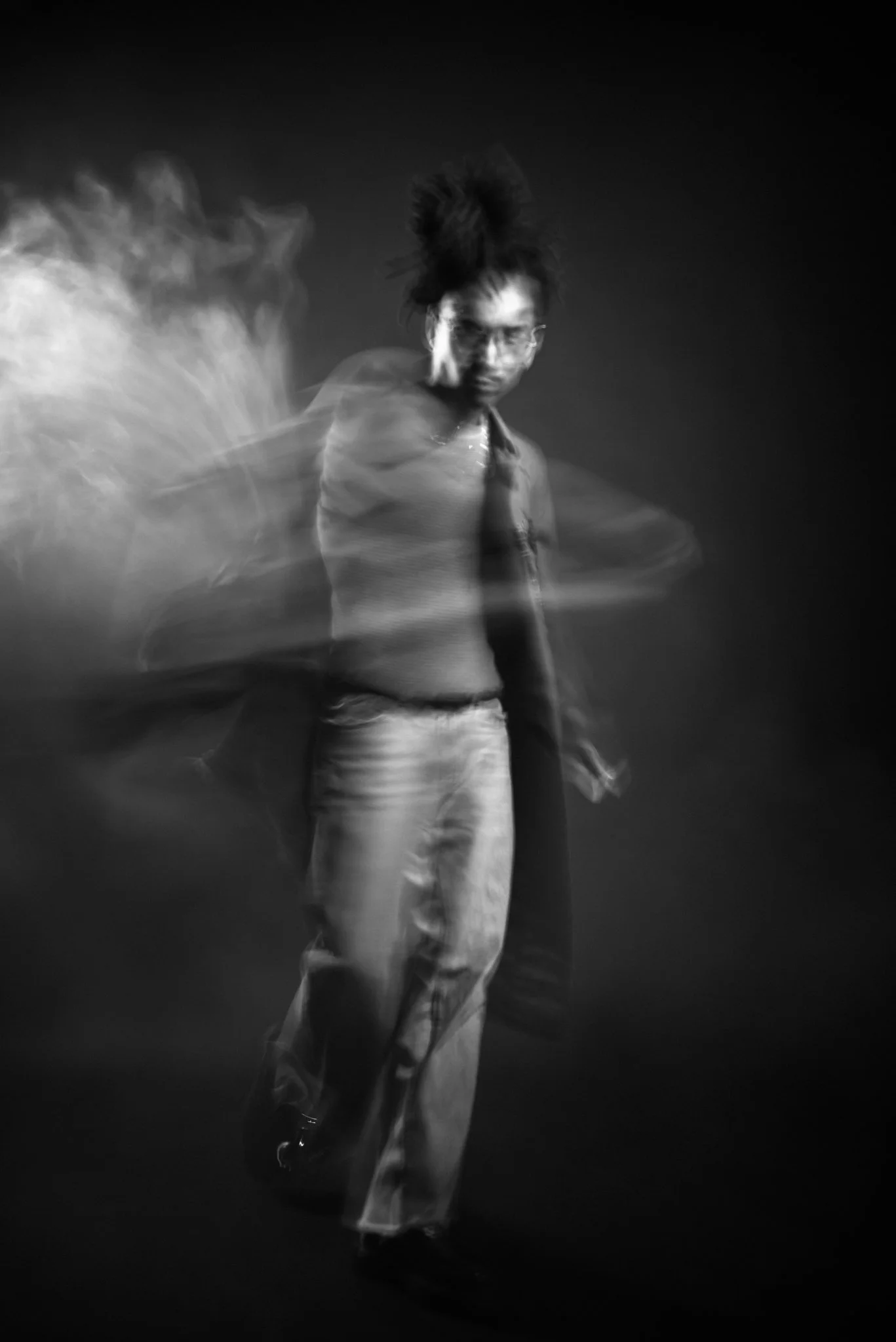 Black and white photo of a person with glasses and dreadlocks, standing against a dark background, with motion blur around them.