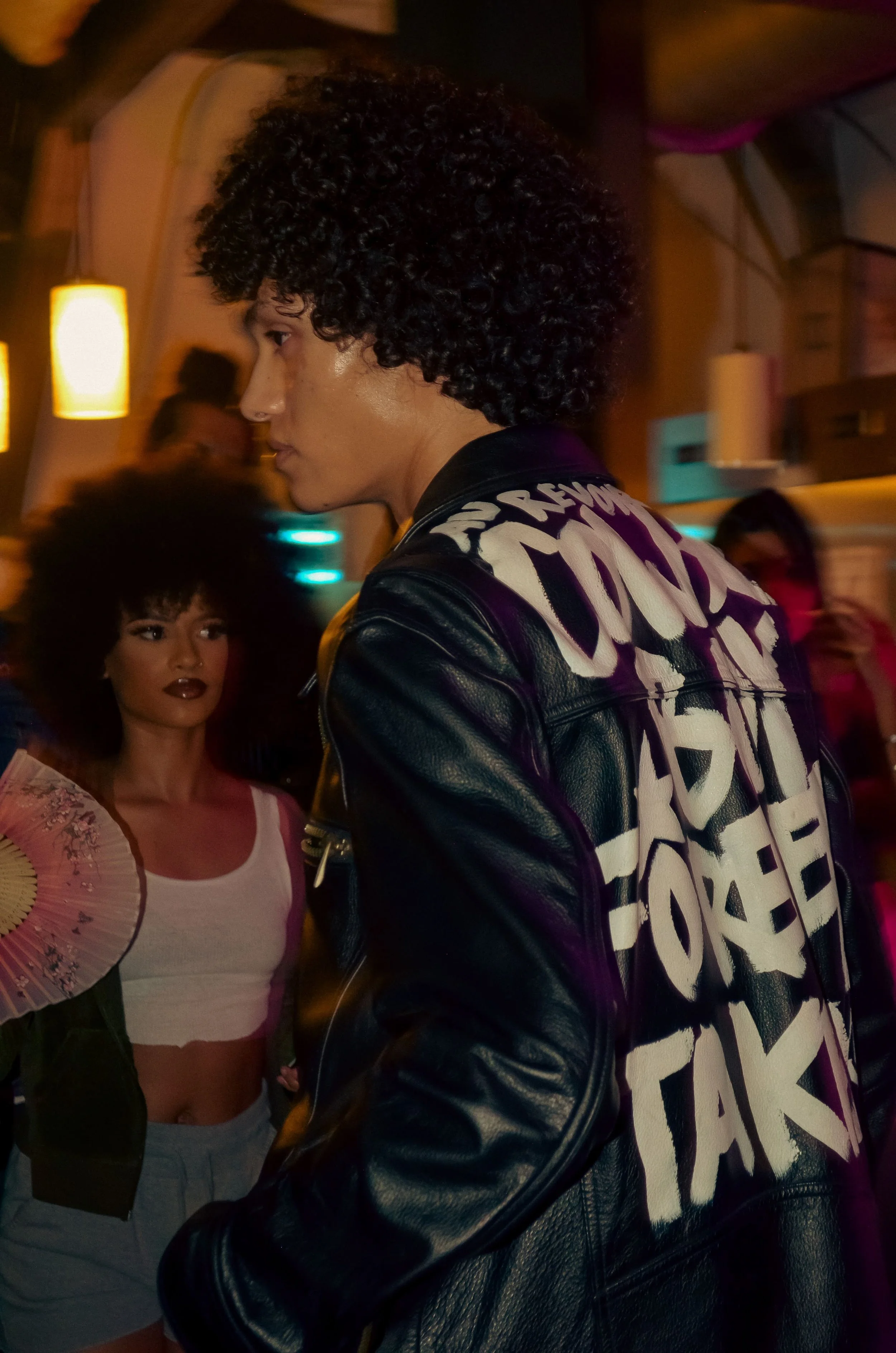 Two women in a dimly lit nightclub or bar. One in the foreground wearing a black leather jacket with bold white writing on the back and curly dark hair. The other in the background holding a fan with curly hair and wearing a white crop top and gray shorts.