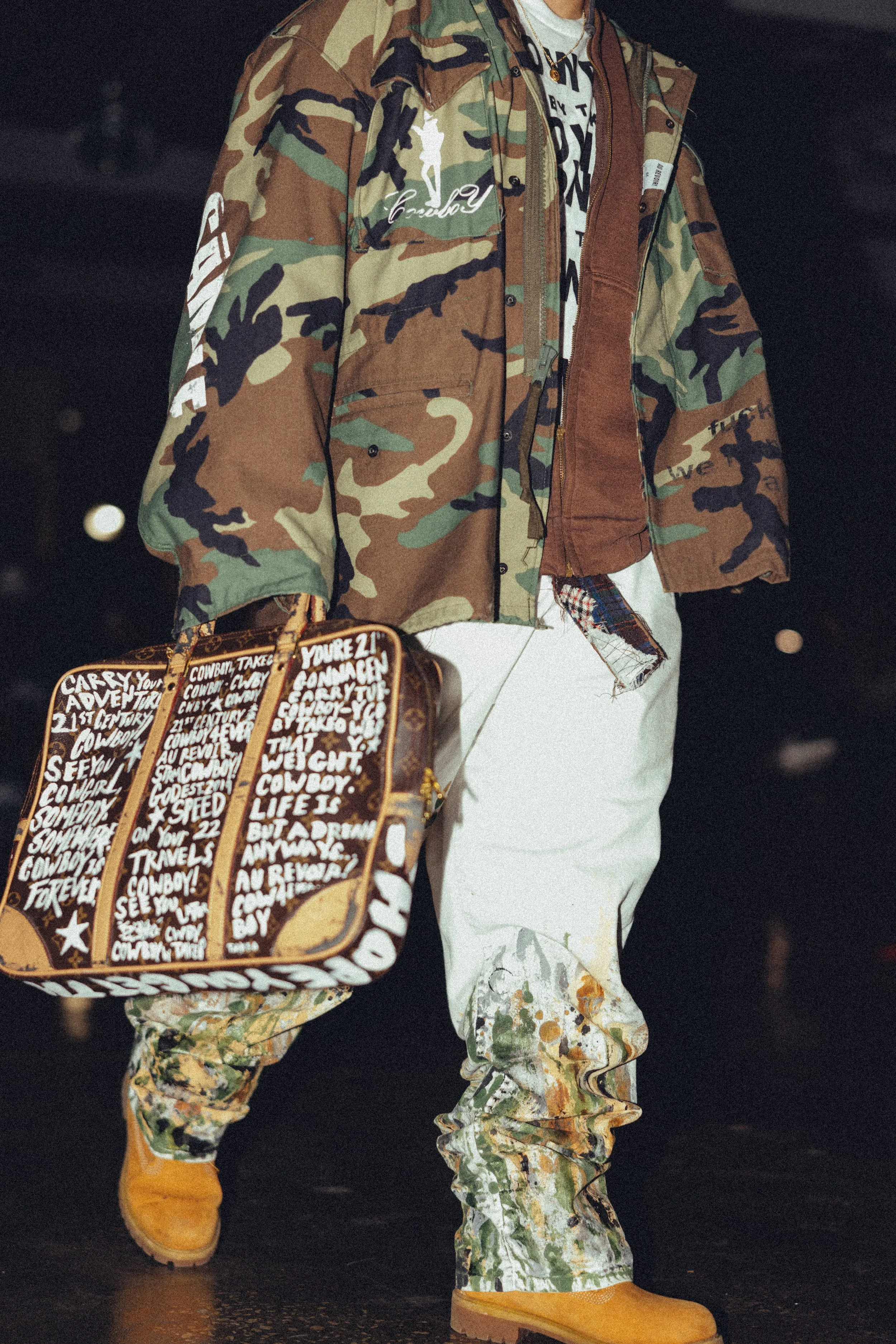 Model walking on a runway wearing a camouflage jacket, white pants with floral patterns, and yellow boots, carrying a brown bag with handwritten text.
