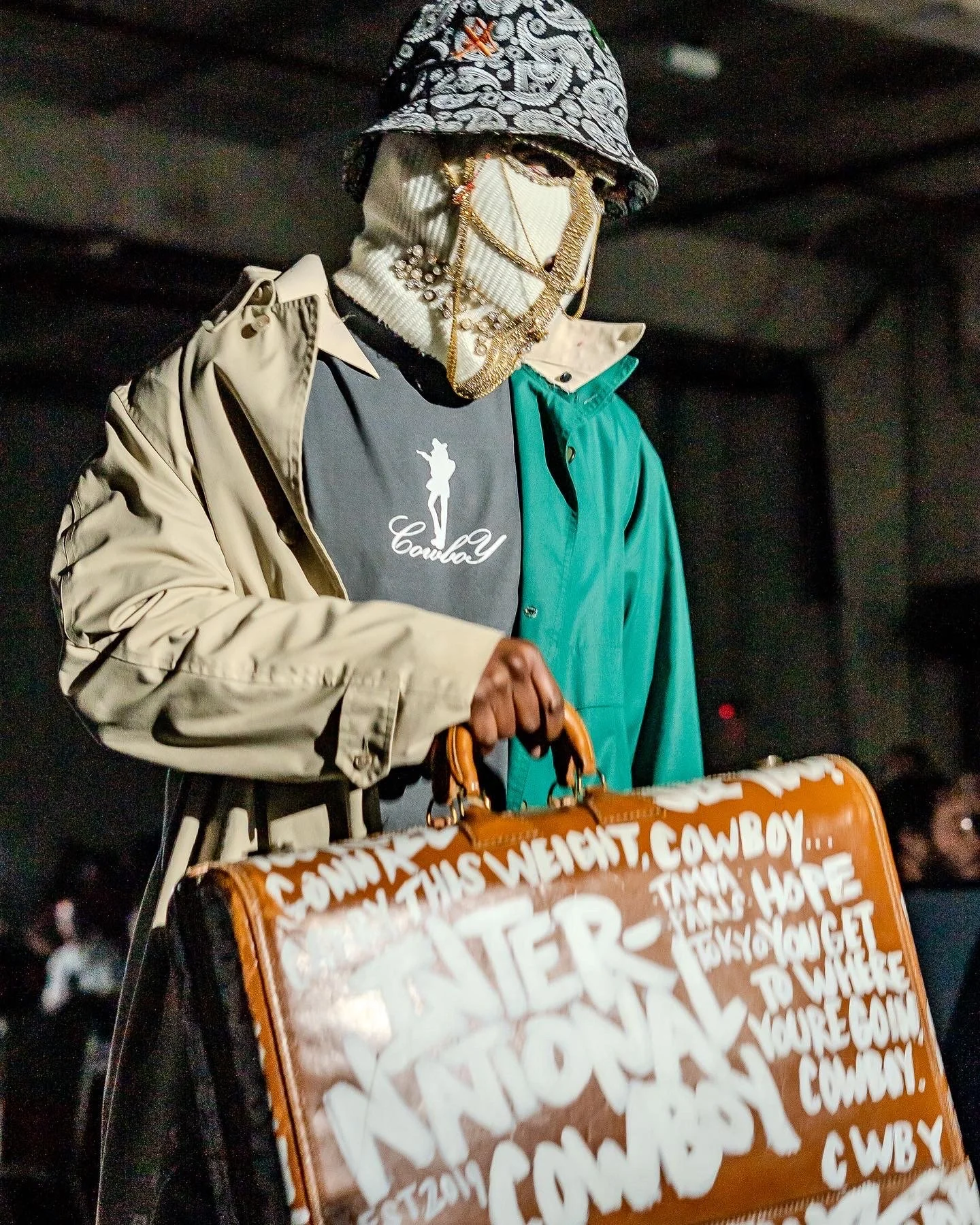 A mannequin dressed in a patterned hat, gold jewelry, a graphic T-shirt, a beige and green jacket, and holding a brown suitcase with white handwritten slogans and white lettering.