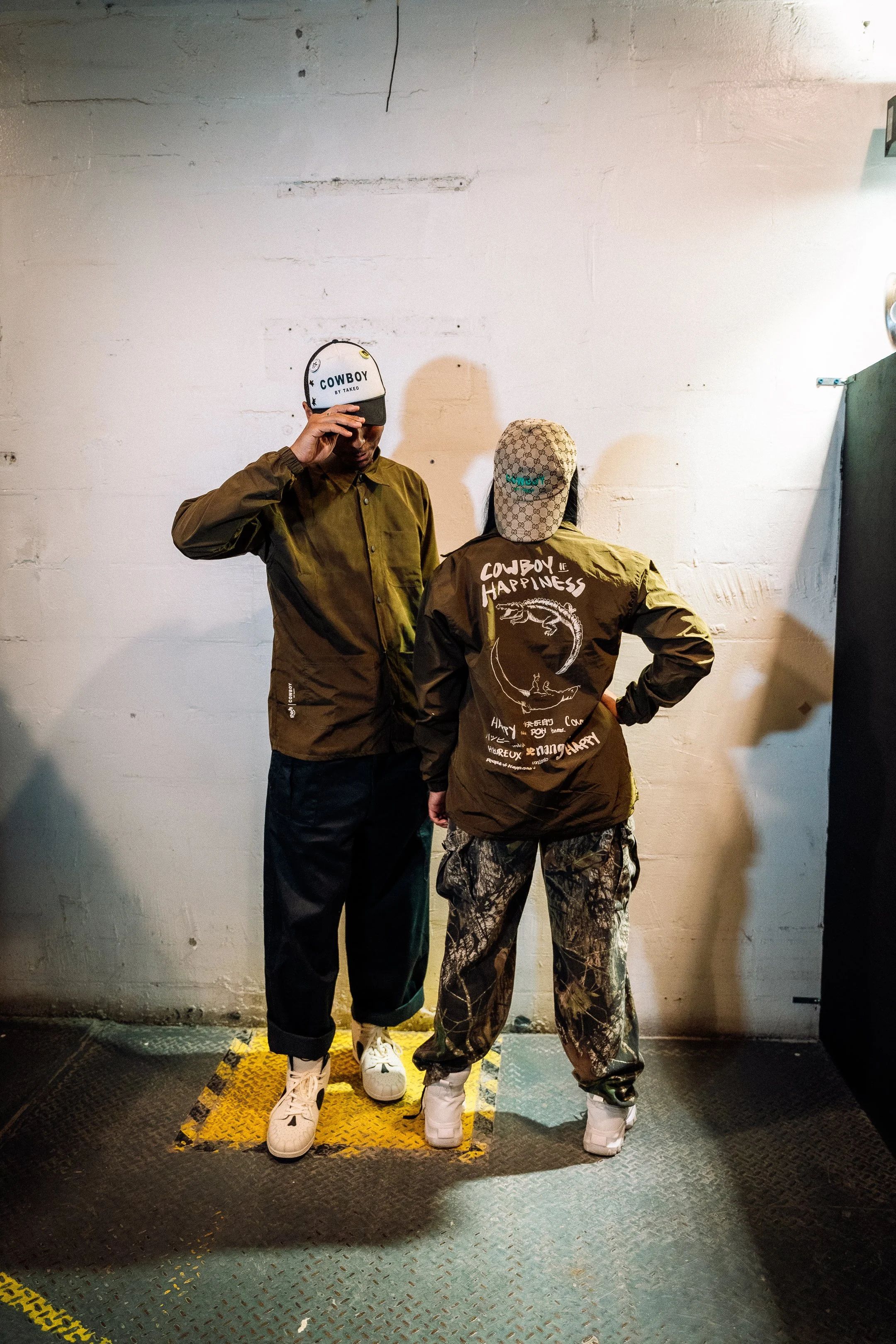Two people standing on a yellow and black platform against a white wall. One person wears a white cap and an olive green jacket, and the other wears a patterned cap and a jacket with "Cowboy Happiness" written on the back. Both wear sneakers.