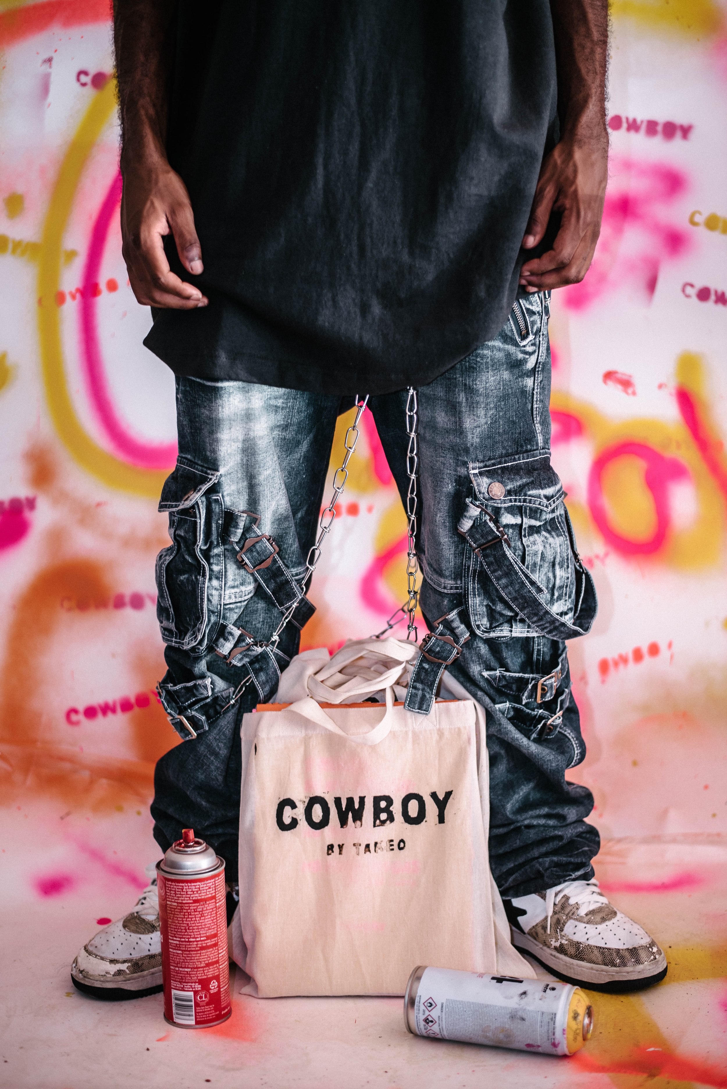 Person standing in front of colorful, graffiti-style background, wearing black oversized shirt, distressed cargo jeans with chains, holding a beige tote bag with the word 'COWBOY' printed on it, and Sneakers. Spray paint cans are on the ground.