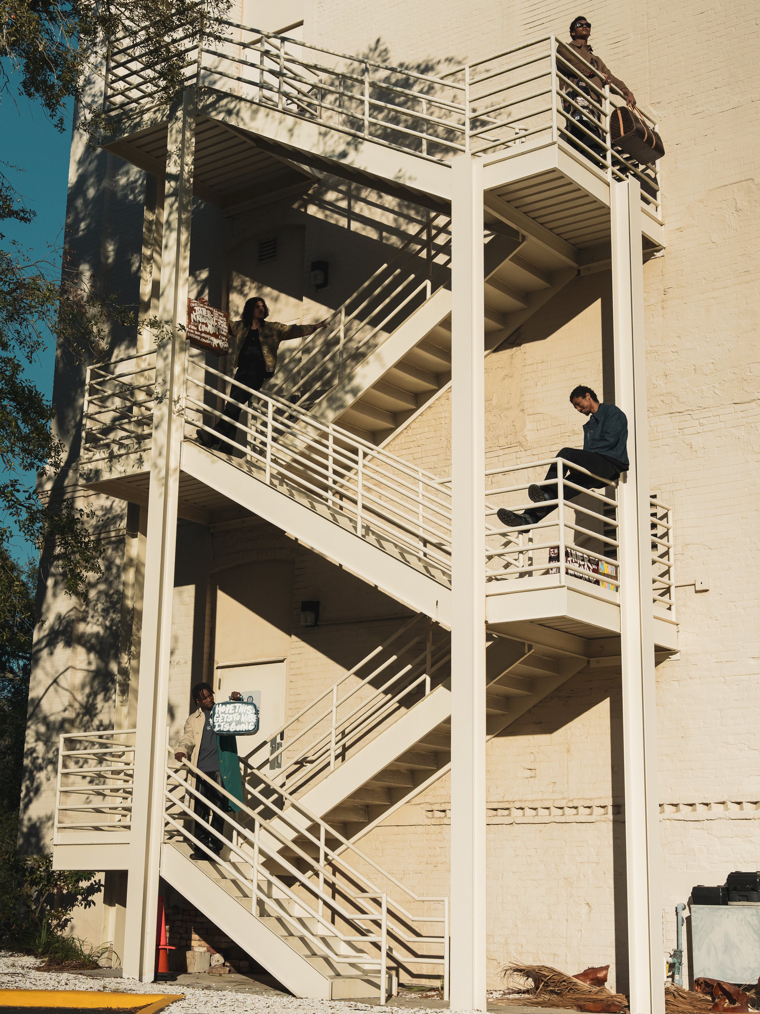 Four people standing or sitting on a white outdoor staircase and landings attached to a building with a brick wall, casting shadows from nearby trees.
