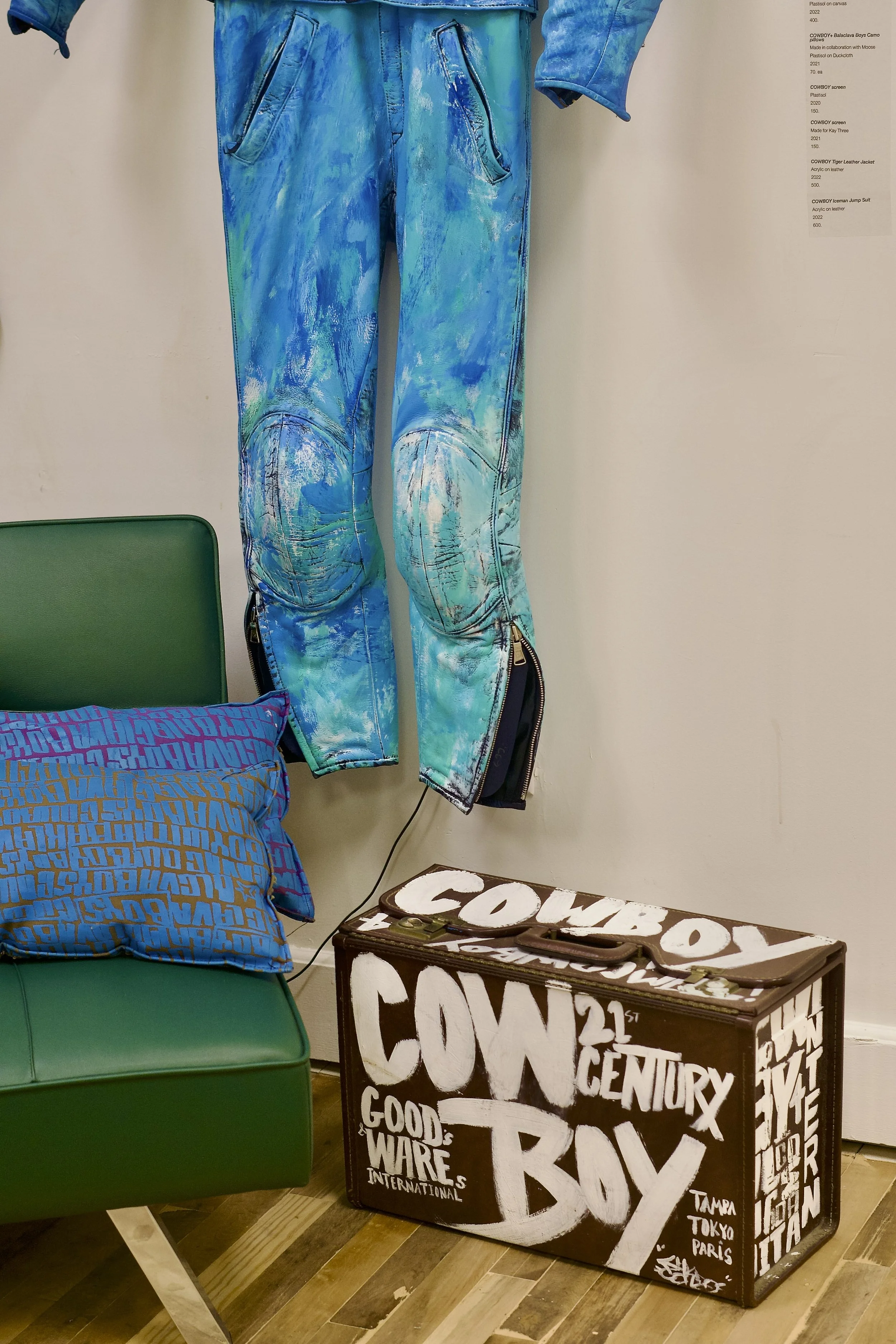 A pair of blue painted jeans hanging on the wall, a green chair with a blue patterned pillow, and a brown box with white lettering that reads 'Cowboy 21st Century Boy' and 'Good Wires International' on the floor.