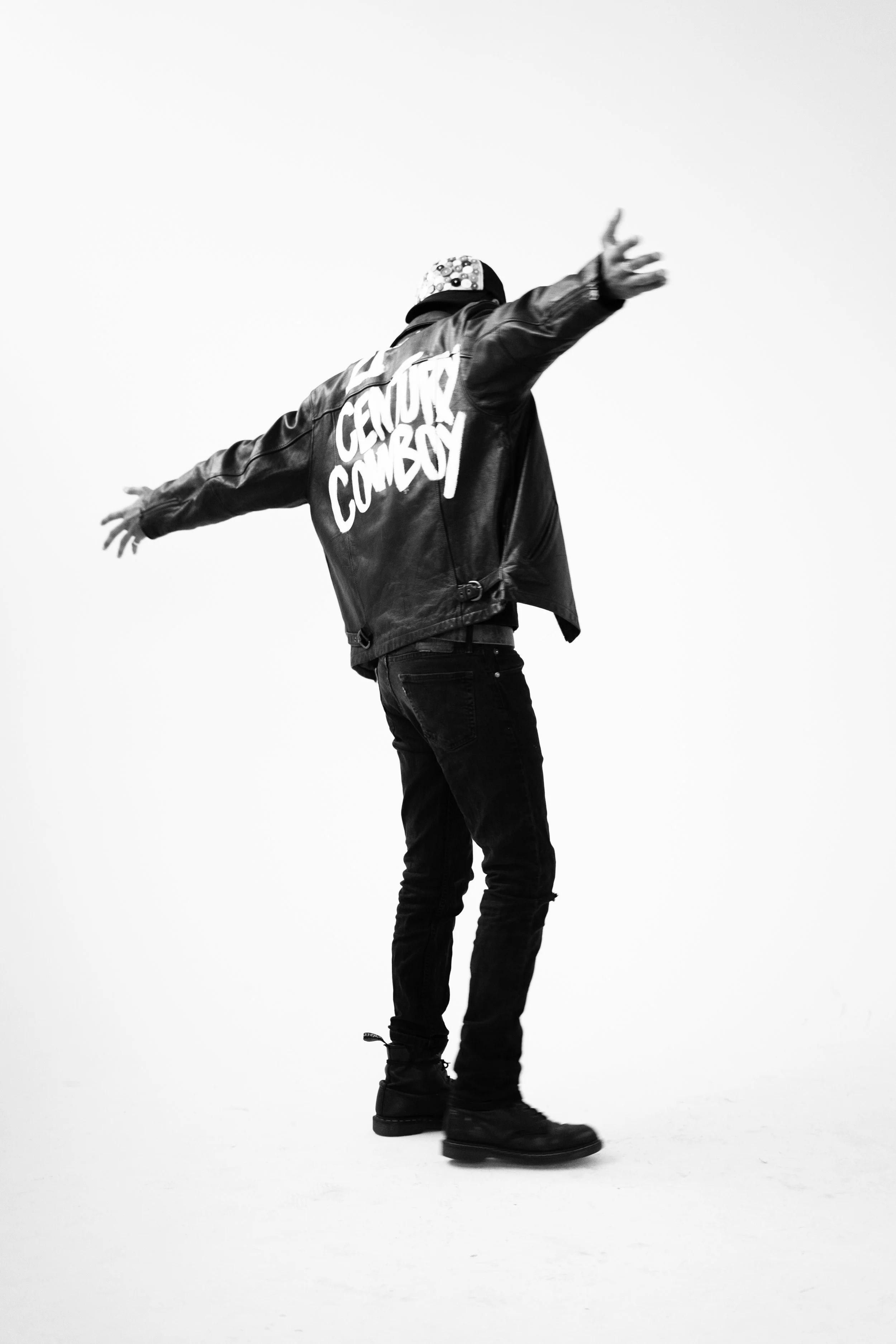A person wearing a leather jacket with 'Genuine Cowboy' written on the back, standing with arms outstretched against a plain background.