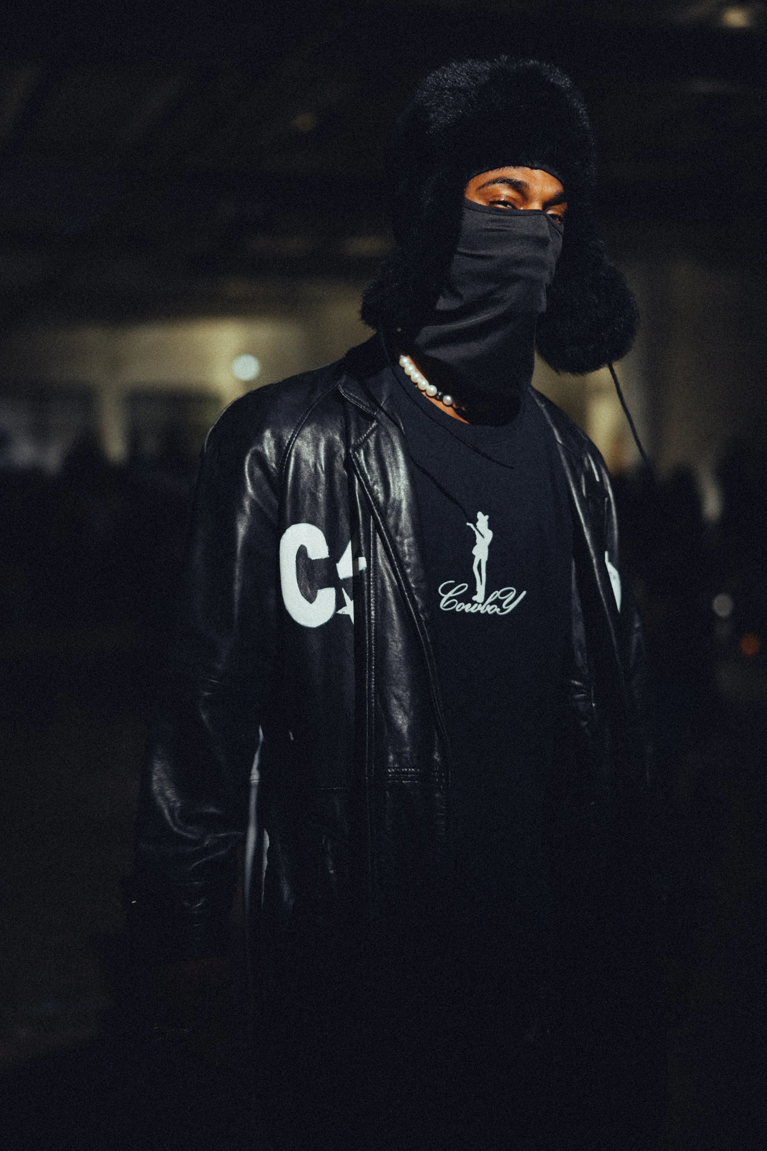 Person wearing a black leather jacket, a black hat with ear flaps, a face mask, and a black t-shirt with white graphics, standing in a dimly lit indoor setting.