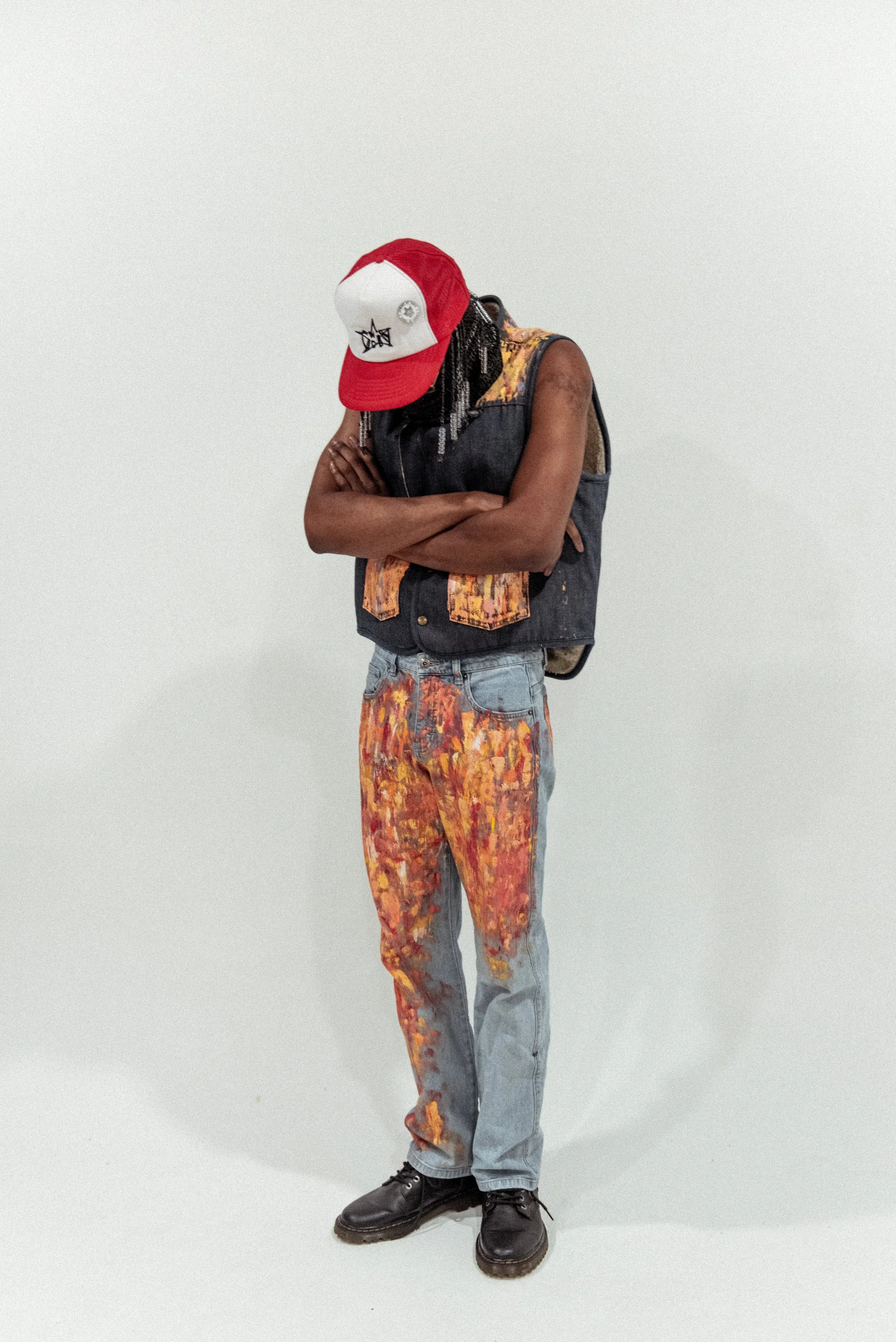 Person standing with arms crossed, wearing a red and white cap, sleeveless denim vest with orange paint splatters, jeans with orange and yellow paint splatters, and black shoes, against a plain white background.