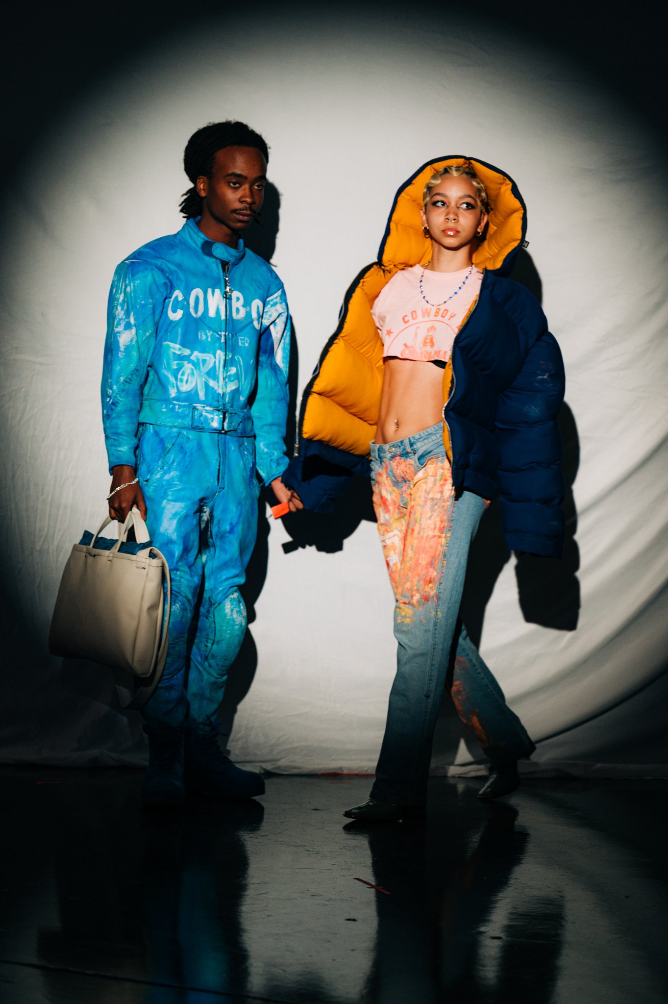 Two models in colorful streetwear pose against a white backdrop. The male model wears a blue jumpsuit with white graffiti and carries a beige bag. The female model wears a yellow hoodie under a black puffer jacket, a pink crop top, and distressed jeans. She accessorizes with a beaded necklace and hoop earrings.