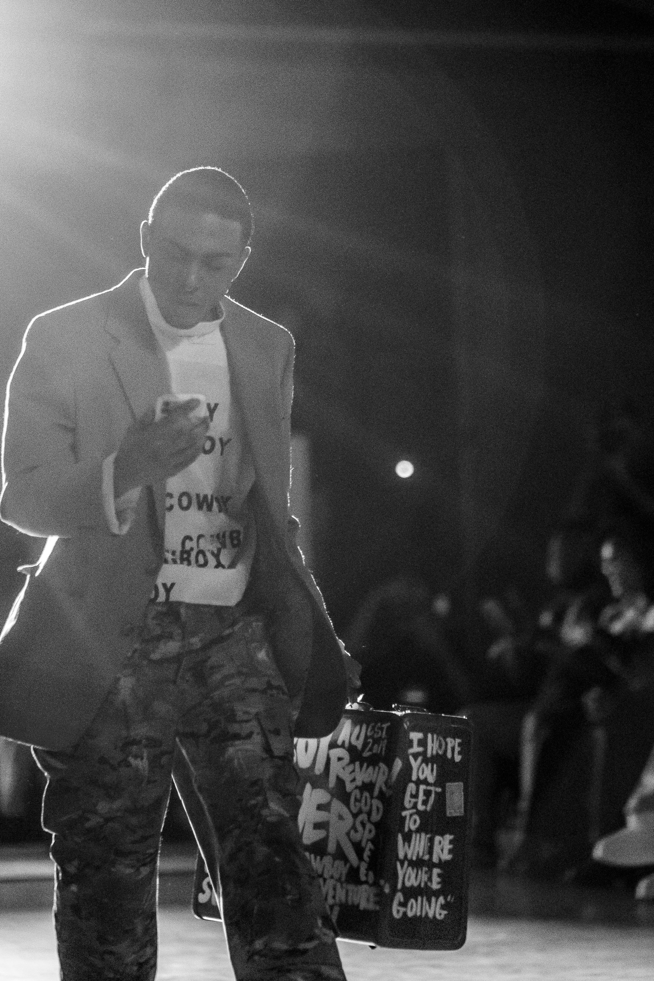 A man wearing a blazer, camouflage pants, and sunglasses is looking at his phone in a dark outdoor setting at night, with a crowd and a sign with handwritten text visible in the background.