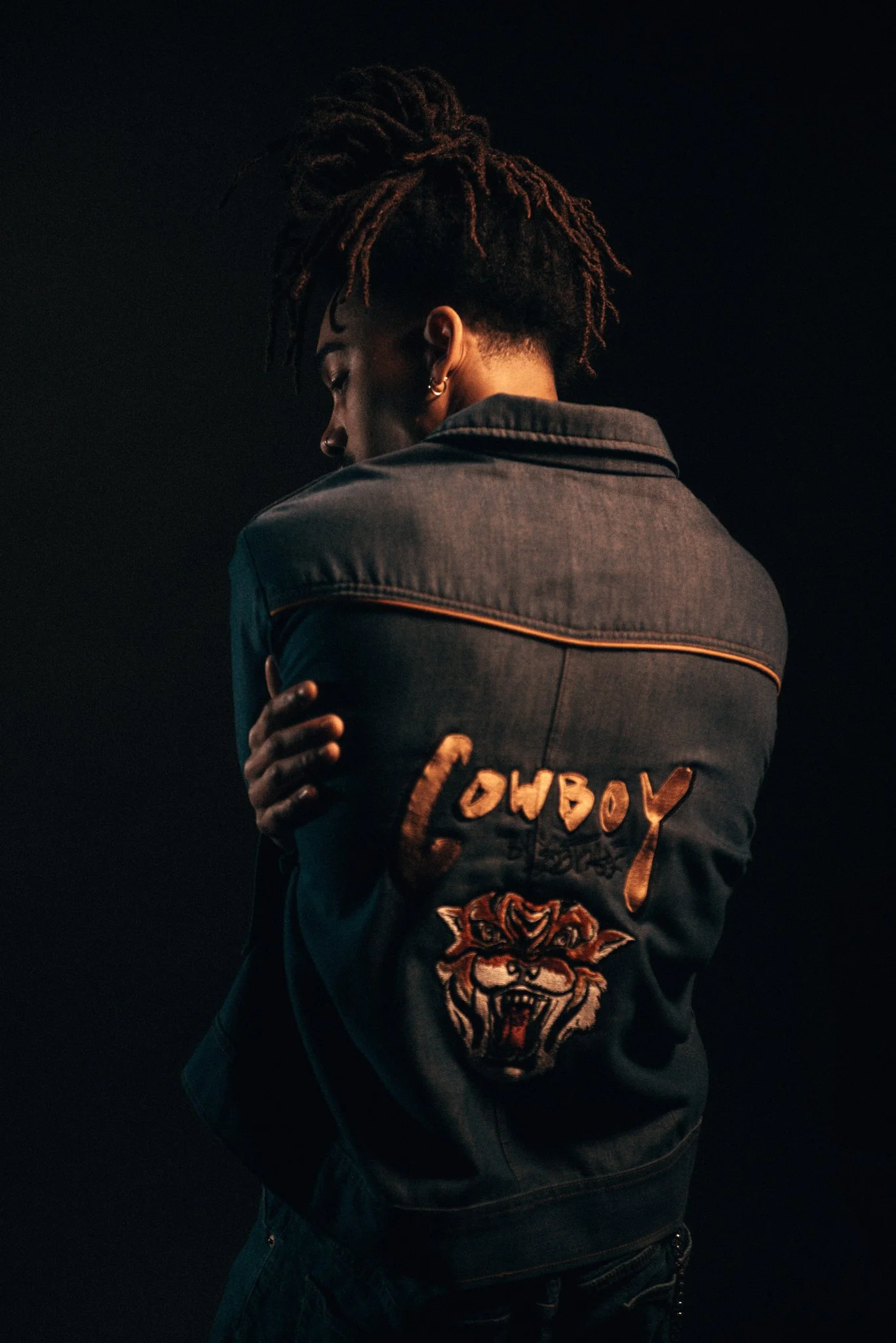 A person with dreadlocks facing away, wearing a denim jacket with embroidered patches that say "LOUBOY" and a fierce tiger face.