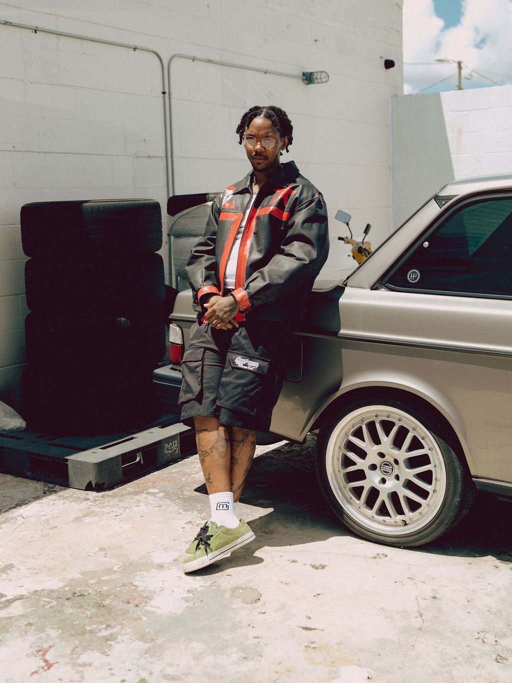 A man leaning against a silver car in an outdoor area, wearing a motorcycle jacket, shorts, sneakers, and glasses, with tires stacked nearby and a motorcycle in the background.