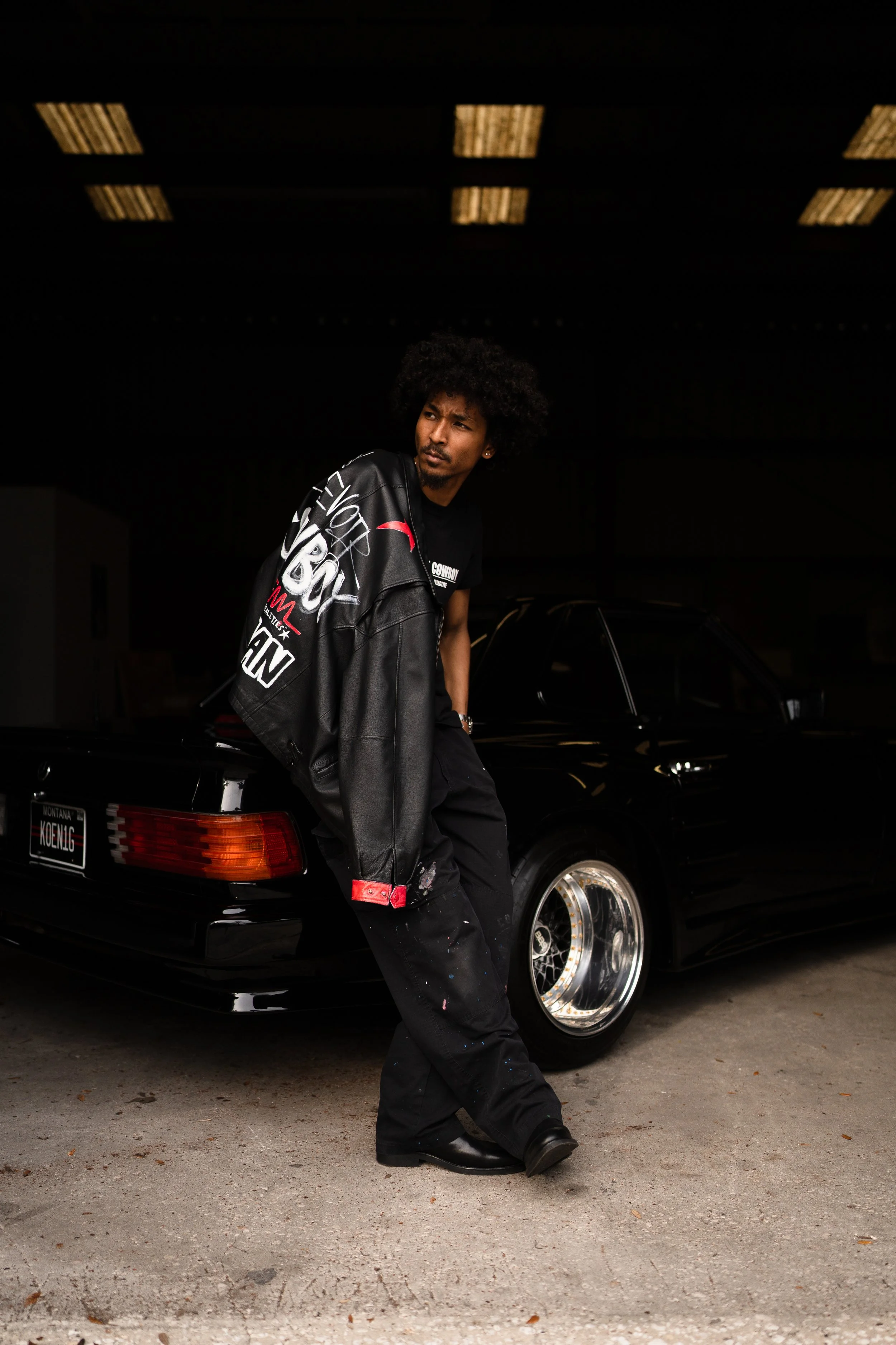 A man with curly hair leaning against a black sports car in an indoor setting with a dark background and overhead lights.