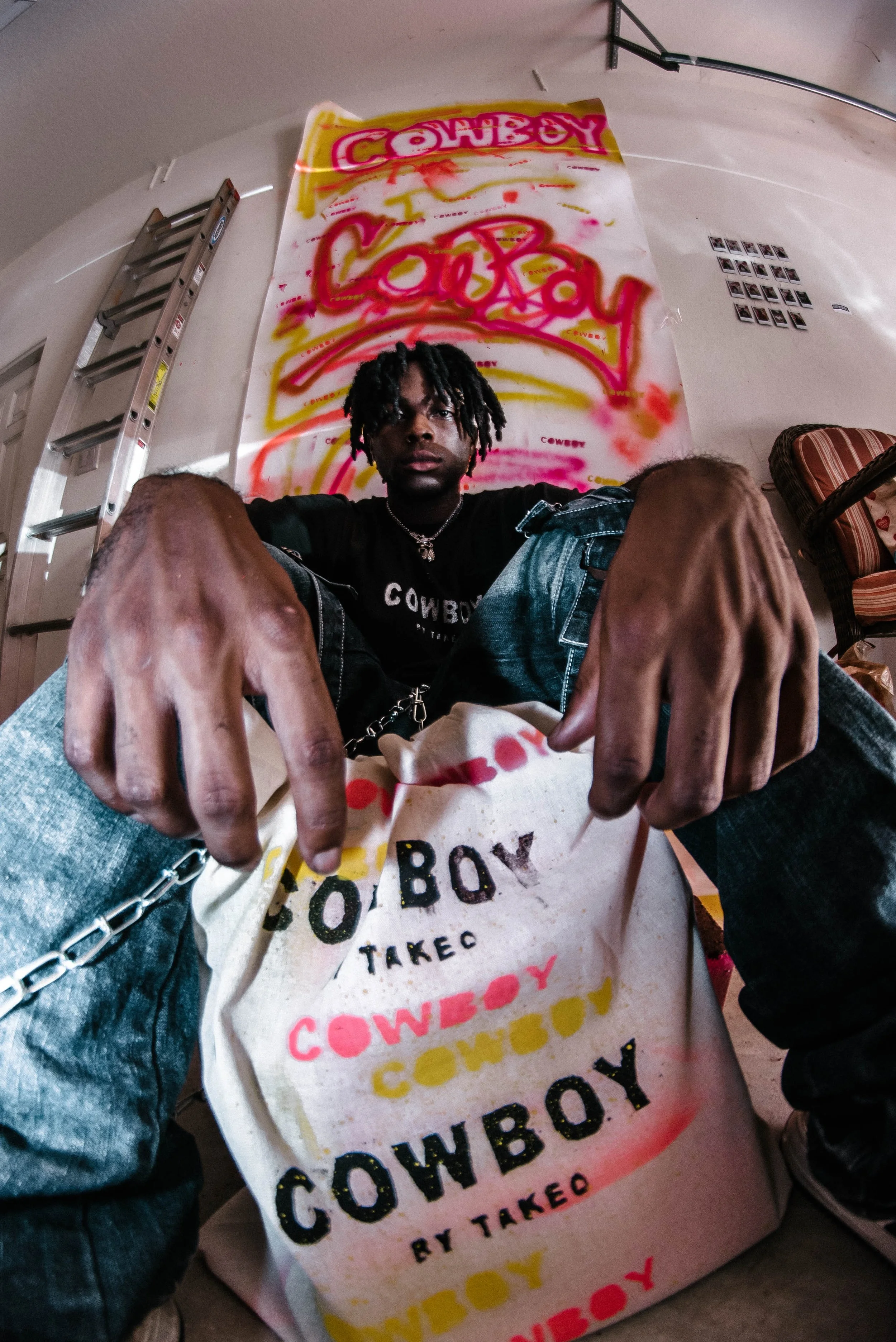 Young man with dreadlocks wearing a black t-shirt and jeans, sitting on the floor, holding a large printed canvas or poster with the words 'COWBOY' and 'TAKEO' in colorful spray paint style, in front of a wall with graffiti art.