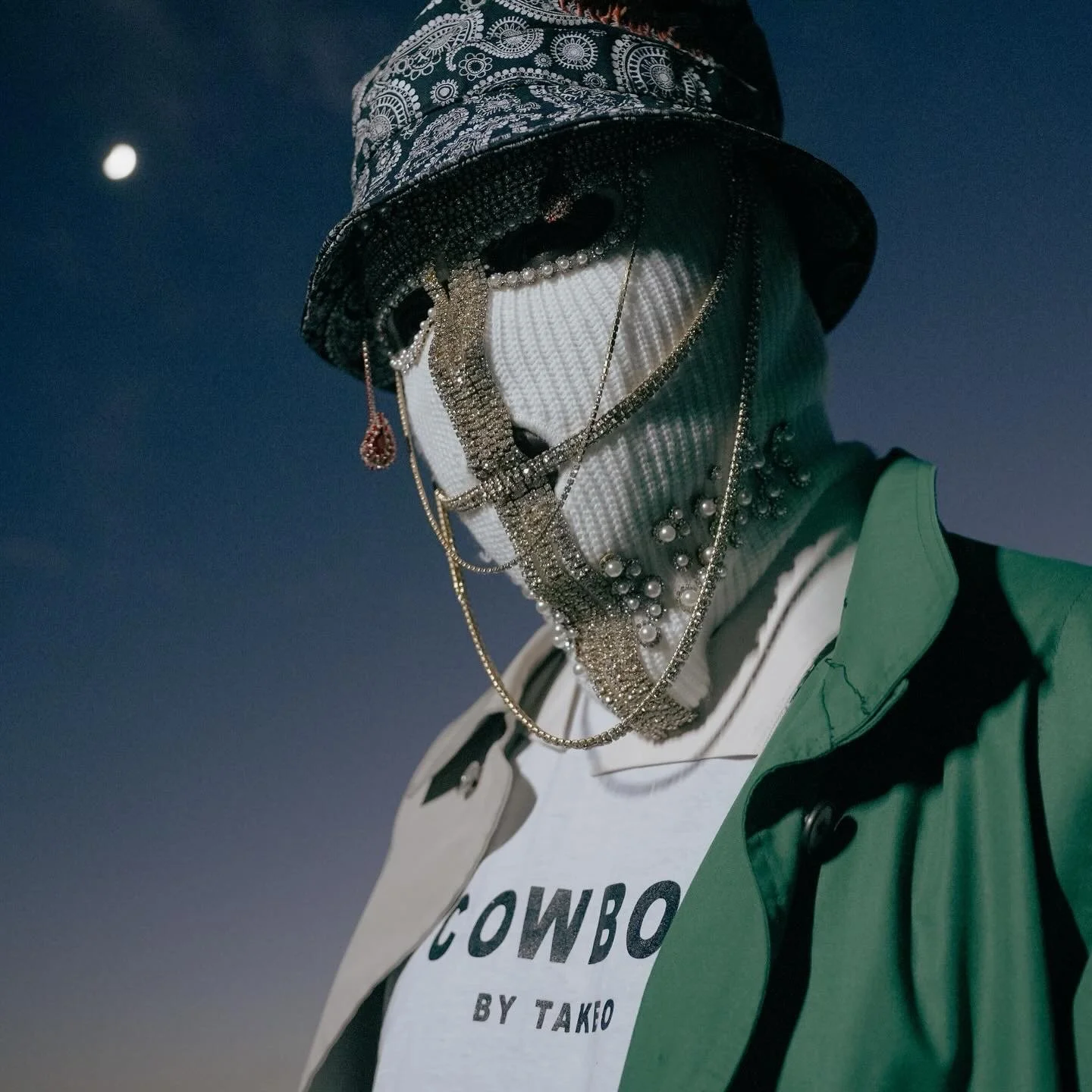 A person wearing a bucket hat and jewelry, with a white face mask, a green jacket, and a white t-shirt with "COWBOY BY TAKO" printed on it, against a dark sky with a visible moon.