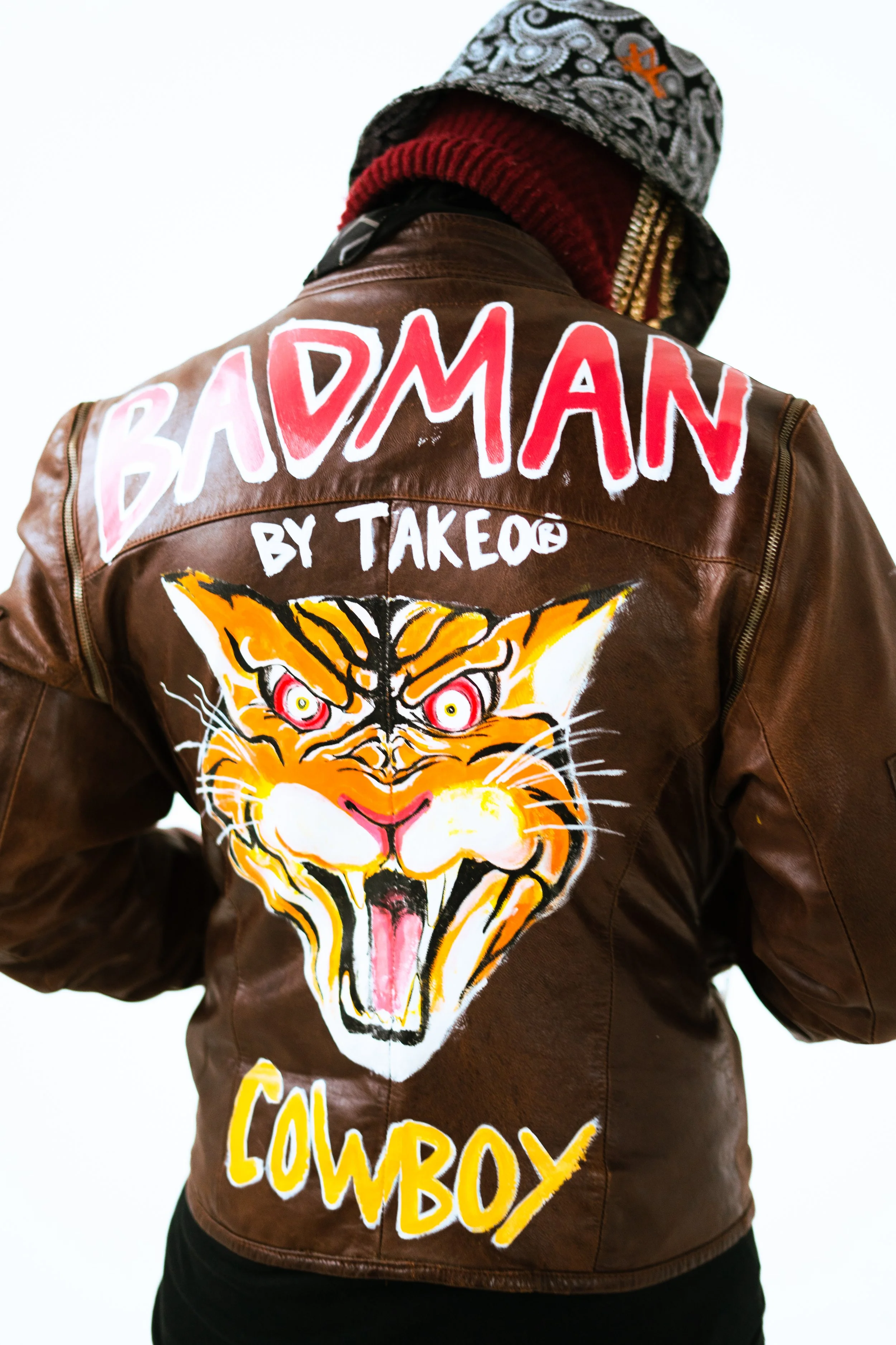 Back of a person wearing a brown leather jacket with colorful text and a tiger graphic. The text reads 'Badman by Takeo' and 'Cowboy.' The tiger has orange, black, and white fur, with pink eyes, yellow accents, and a pink tongue showing.