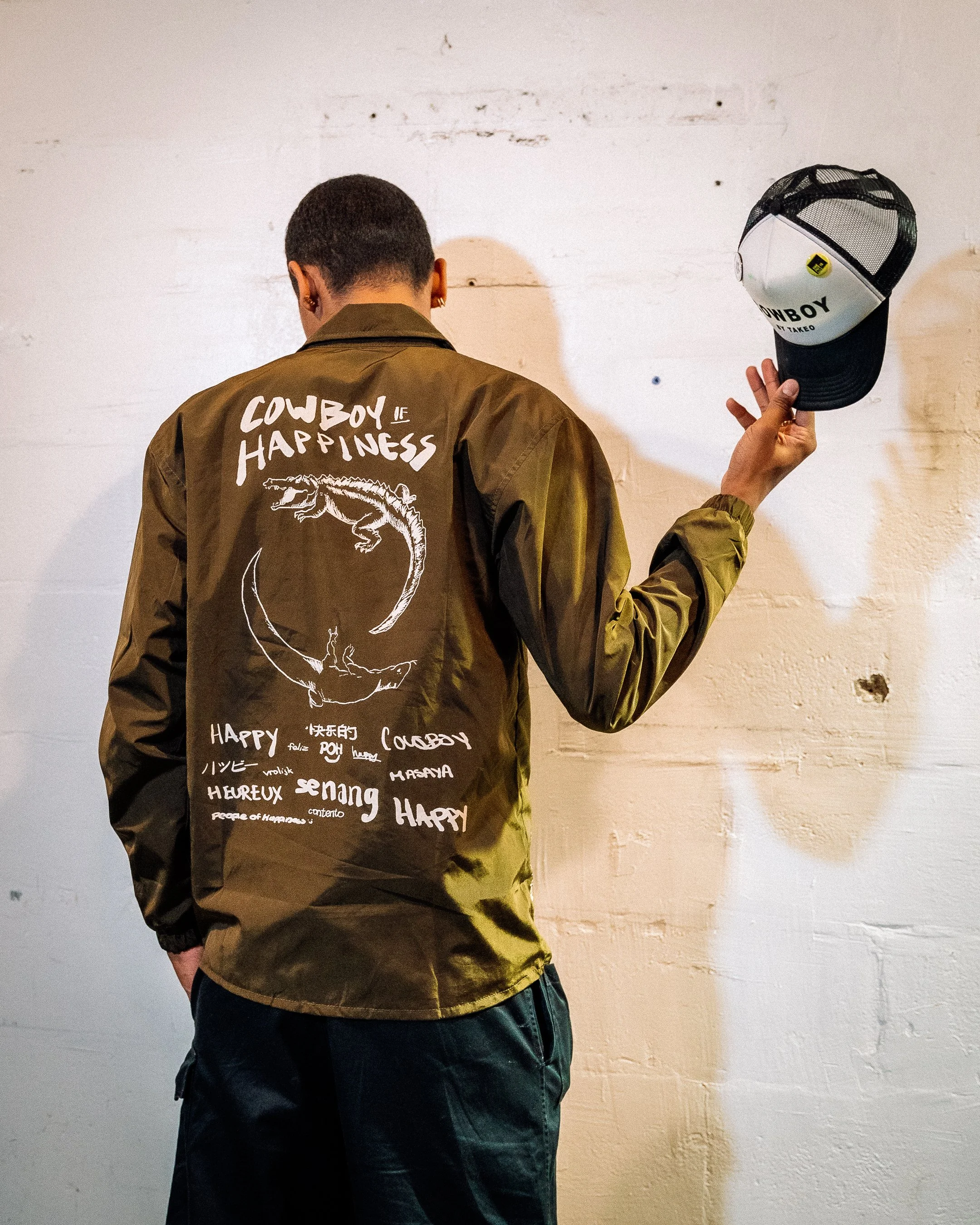 Back of a person wearing a jacket with drawings and words, holding a baseball cap, standing against a light-colored wall.
