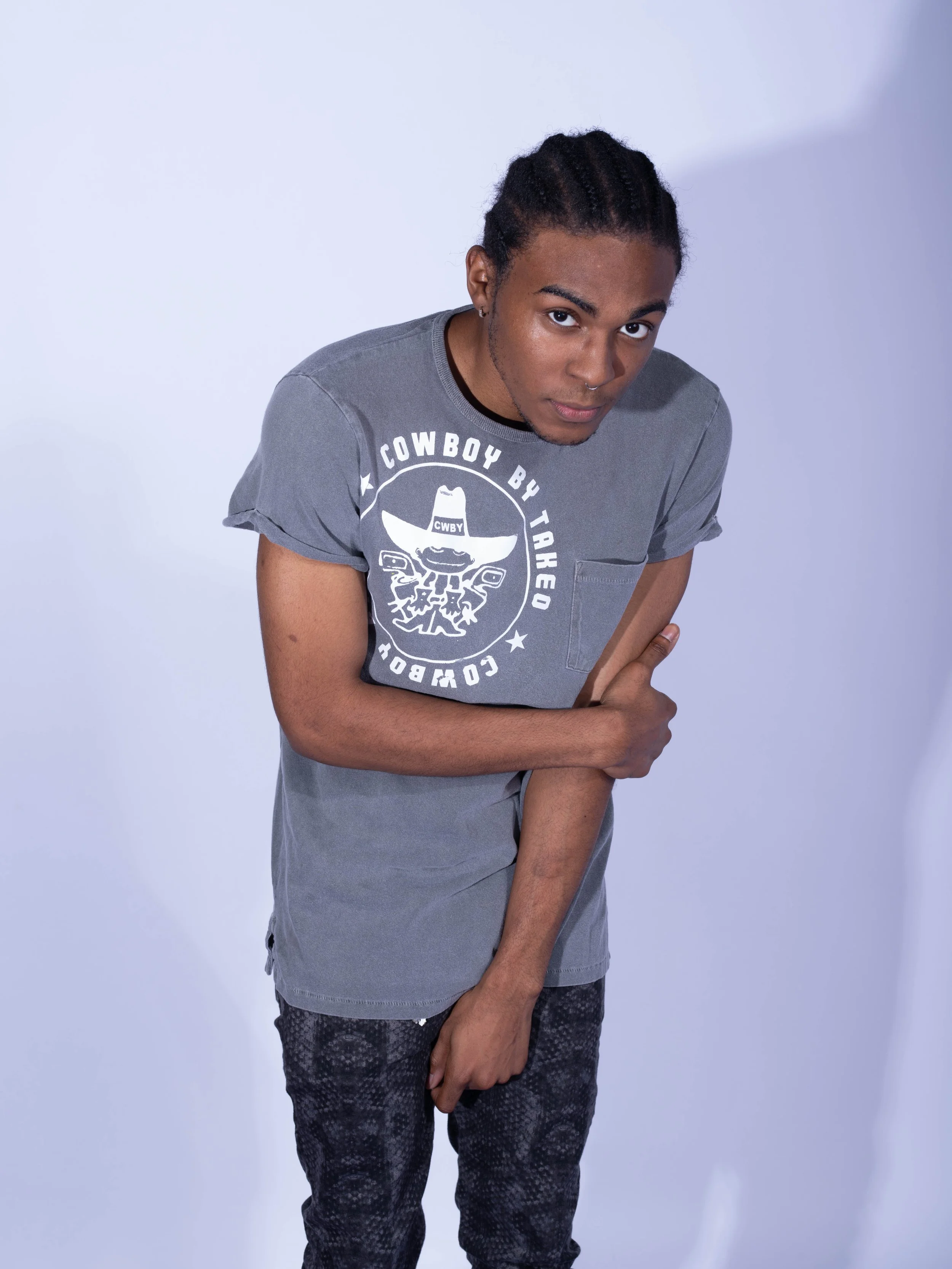 A young man with dark hair in braids, wearing a gray T-shirt with graphic print and dark patterned pants, posing against a white background.
