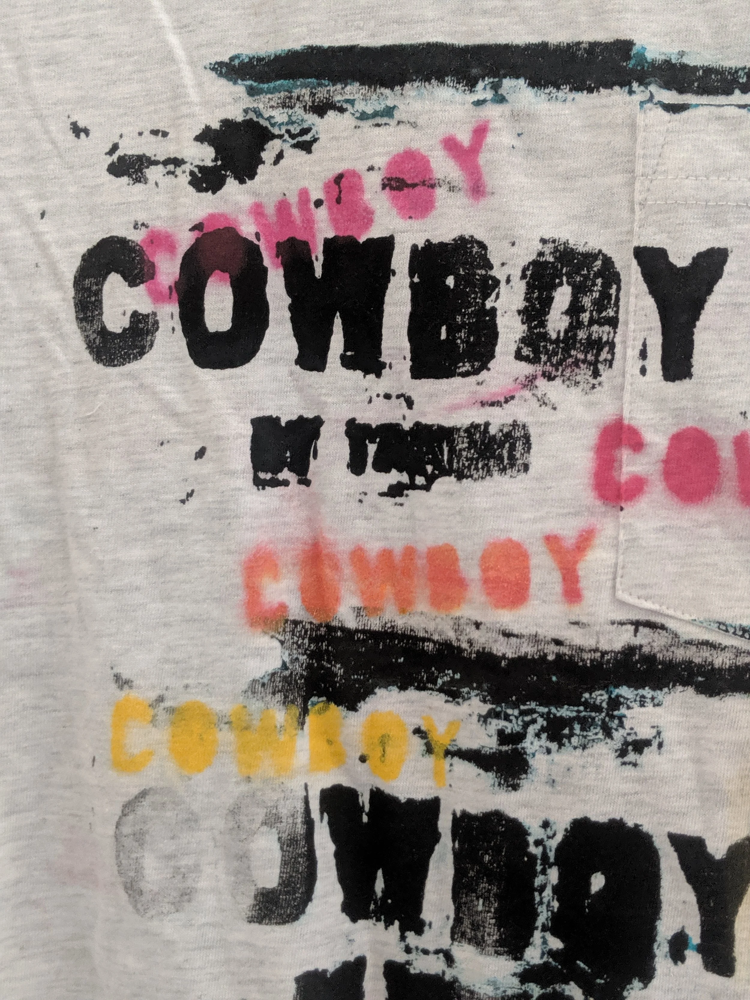 Close-up of a T-shirt with multiple colorful, distressed printed words including 'COWBOY' in black, pink, orange, and yellow, with some letters overlapping or faded.