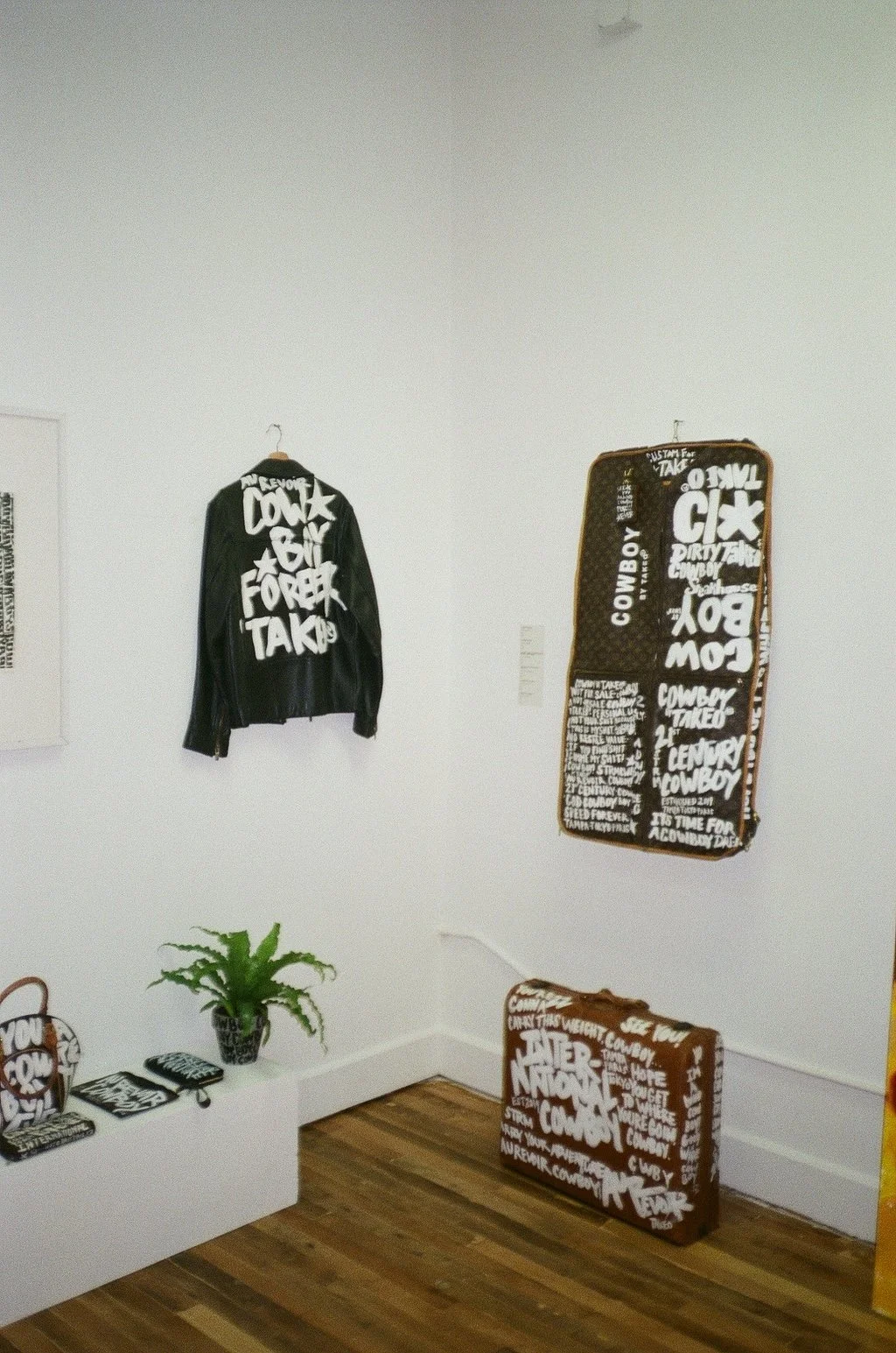 Exhibition of black and brown clothing and accessories with bold white graffiti-style text, a green potted plant, and wooden flooring.