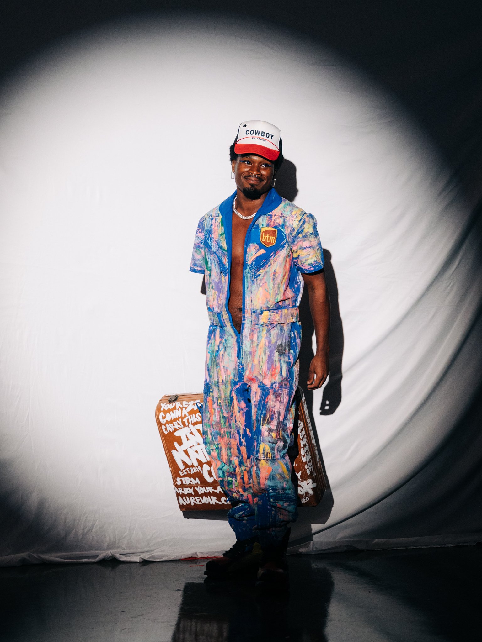 A man standing against a white background, wearing a colorful paint-splattered jumpsuit, a white cap with red, black, and white colors, and carrying a decorated brown suitcase.