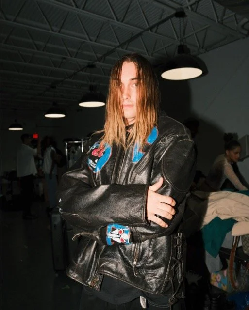 Young woman with shoulder-length hair, wearing a black leather jacket with a colorful patterned hood, standing in a dimly lit indoor space with other people in the background.