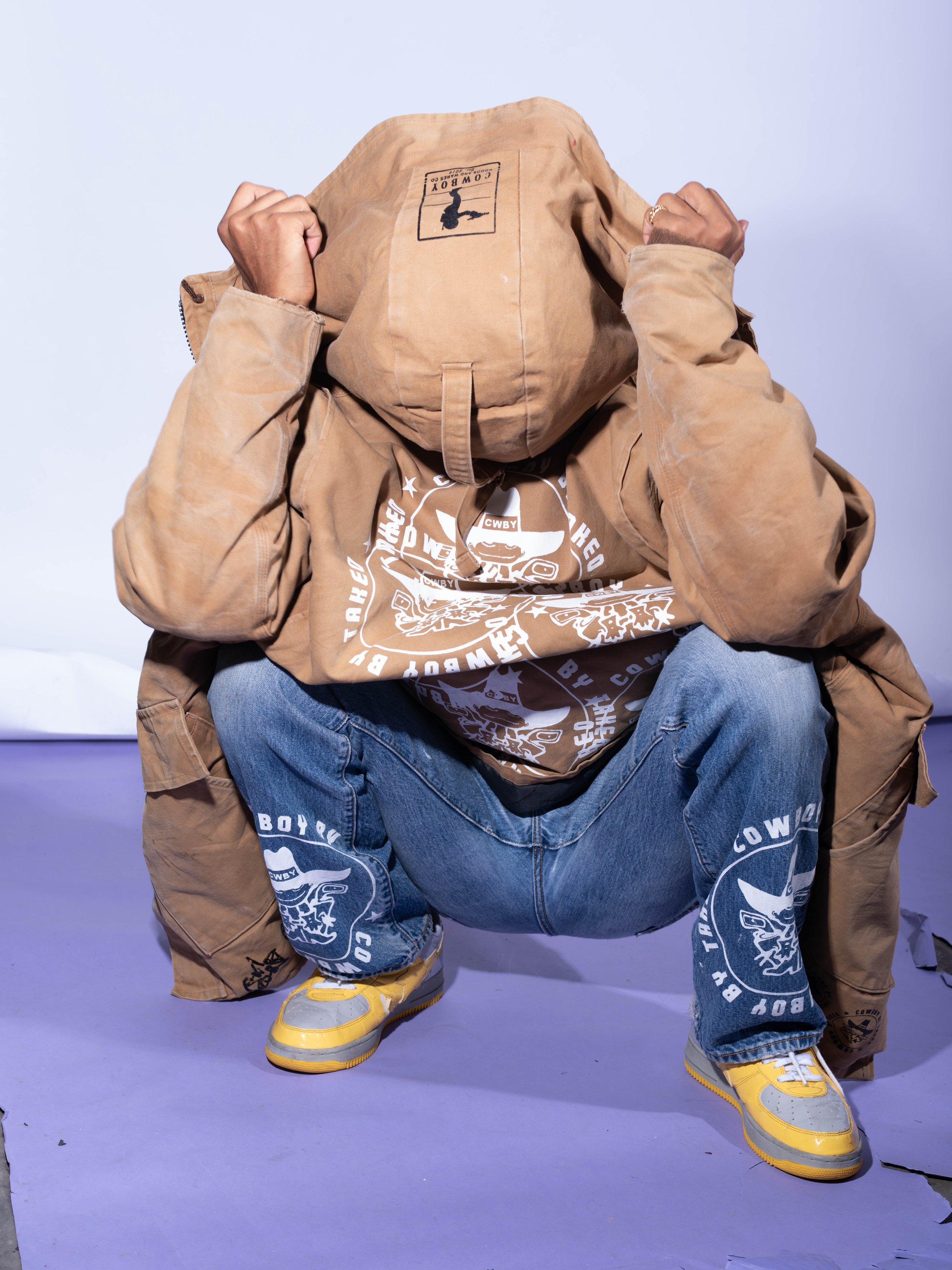 Person crouching with face covered by a tan jacket, wearing blue jeans and yellow sneakers.