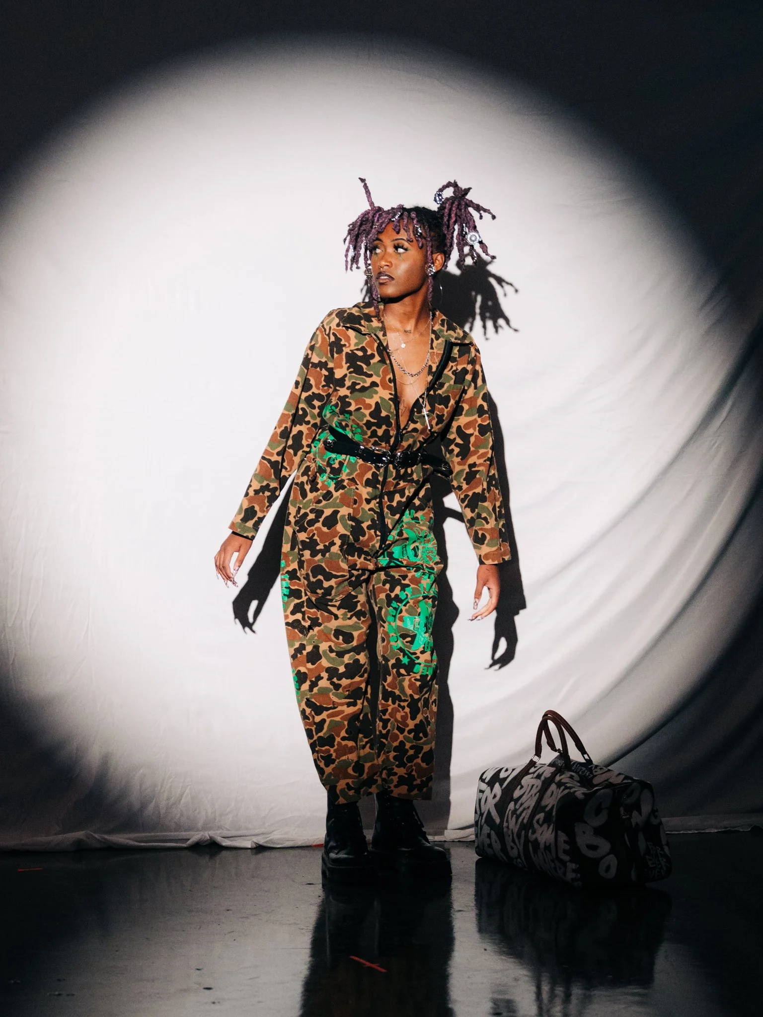 A woman with purple dreadlocks in a camouflage outfit and chunky boots standing on a dark floor, with a duffel bag nearby, under a spotlight.