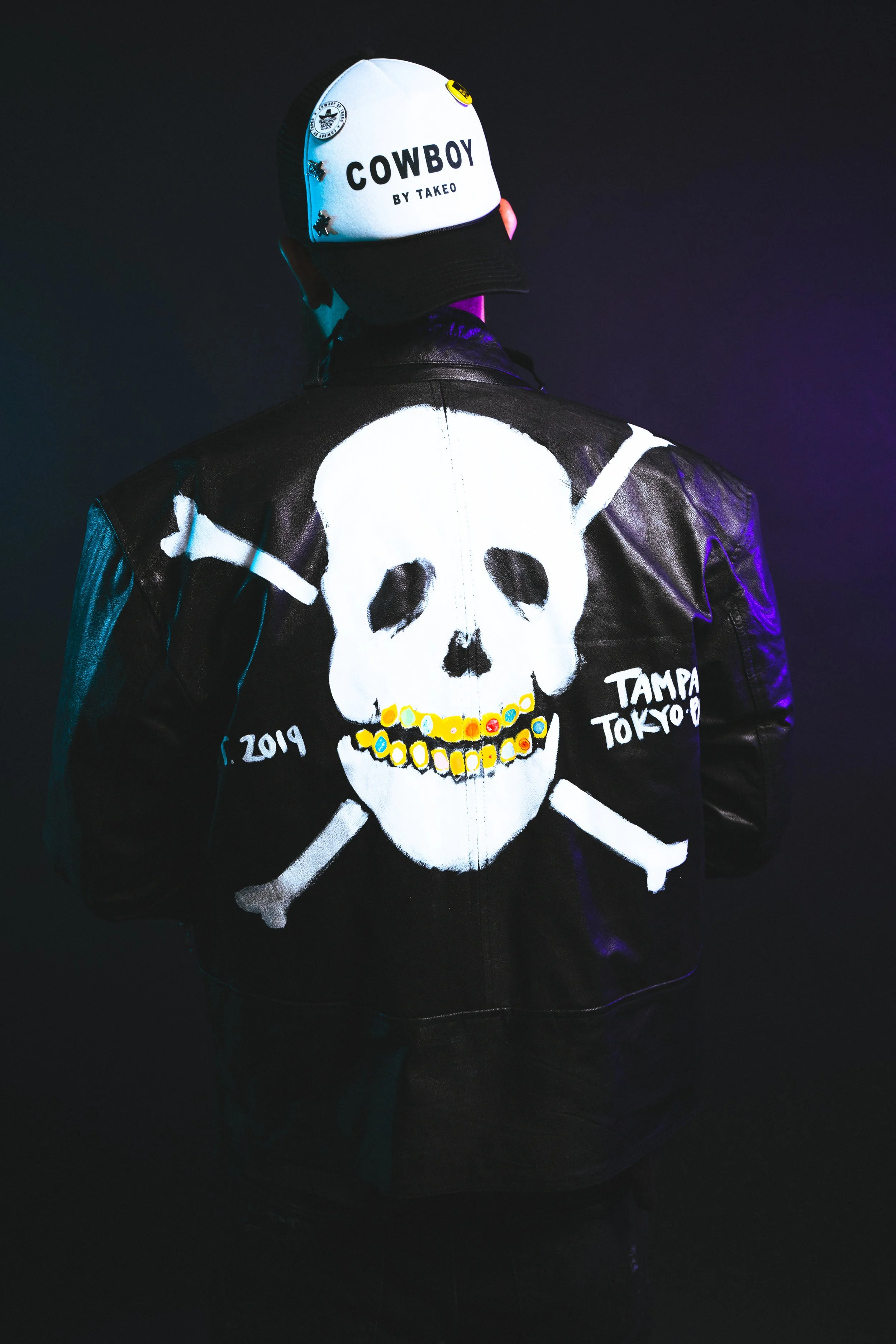Person wearing a black leather jacket with a large white skull and crossbones design on the back, and a white cap with black and yellow patches and the word 'COWBOY BY TAKEO' on it.