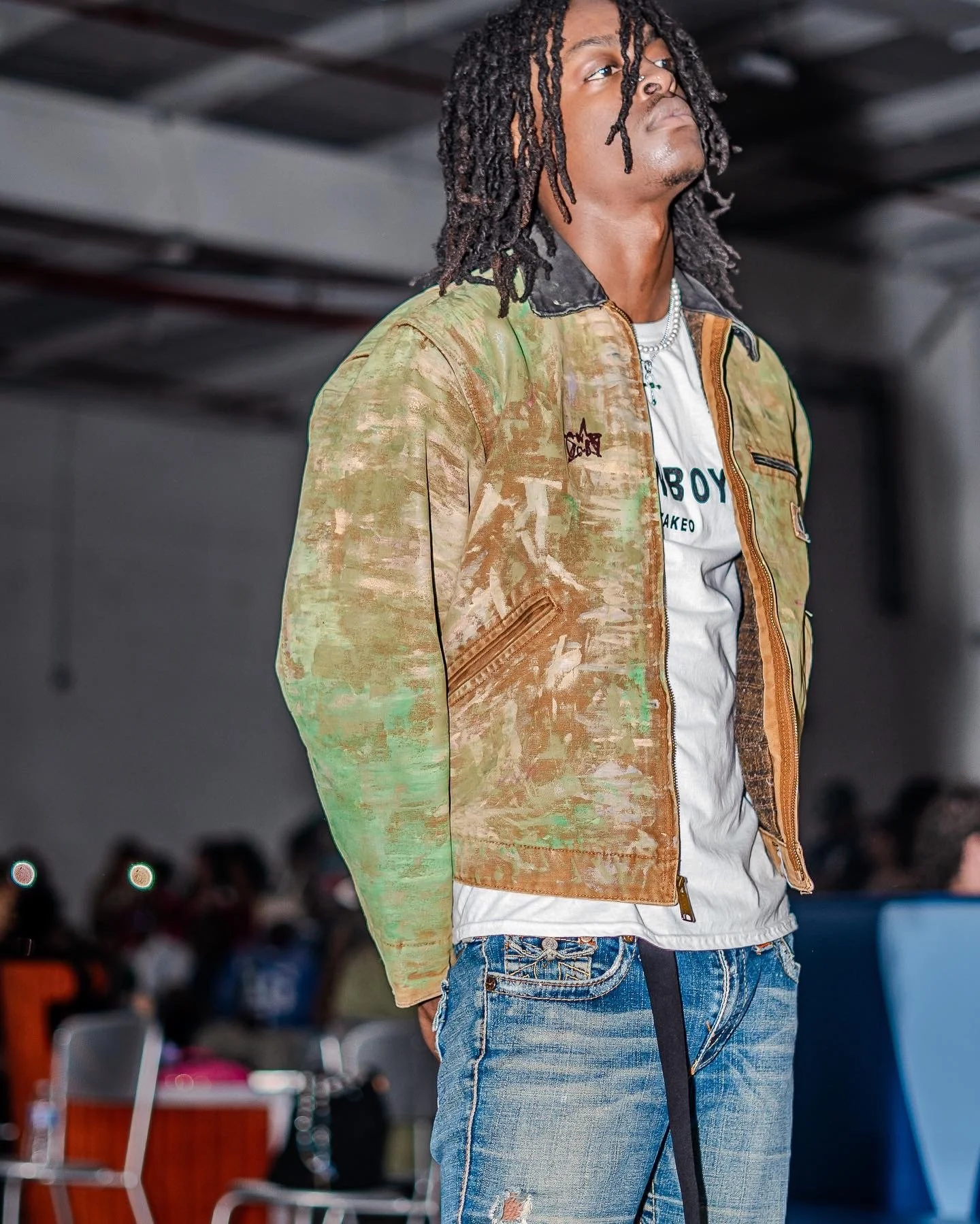 A young man with dreadlocks standing indoors in front of a blurred audience, wearing a colorful tie-dye jacket over a white t-shirt and ripped jeans.