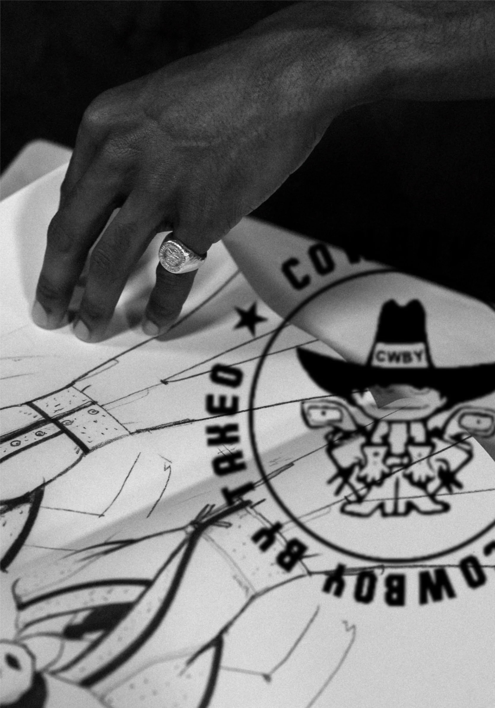 A hand with a ring on the index finger placed on a drawing or diagram, overlaid with a circular logo that features a cartoon character wearing a cowboy hat and the text "COWBOY CHECK ARCHDE".