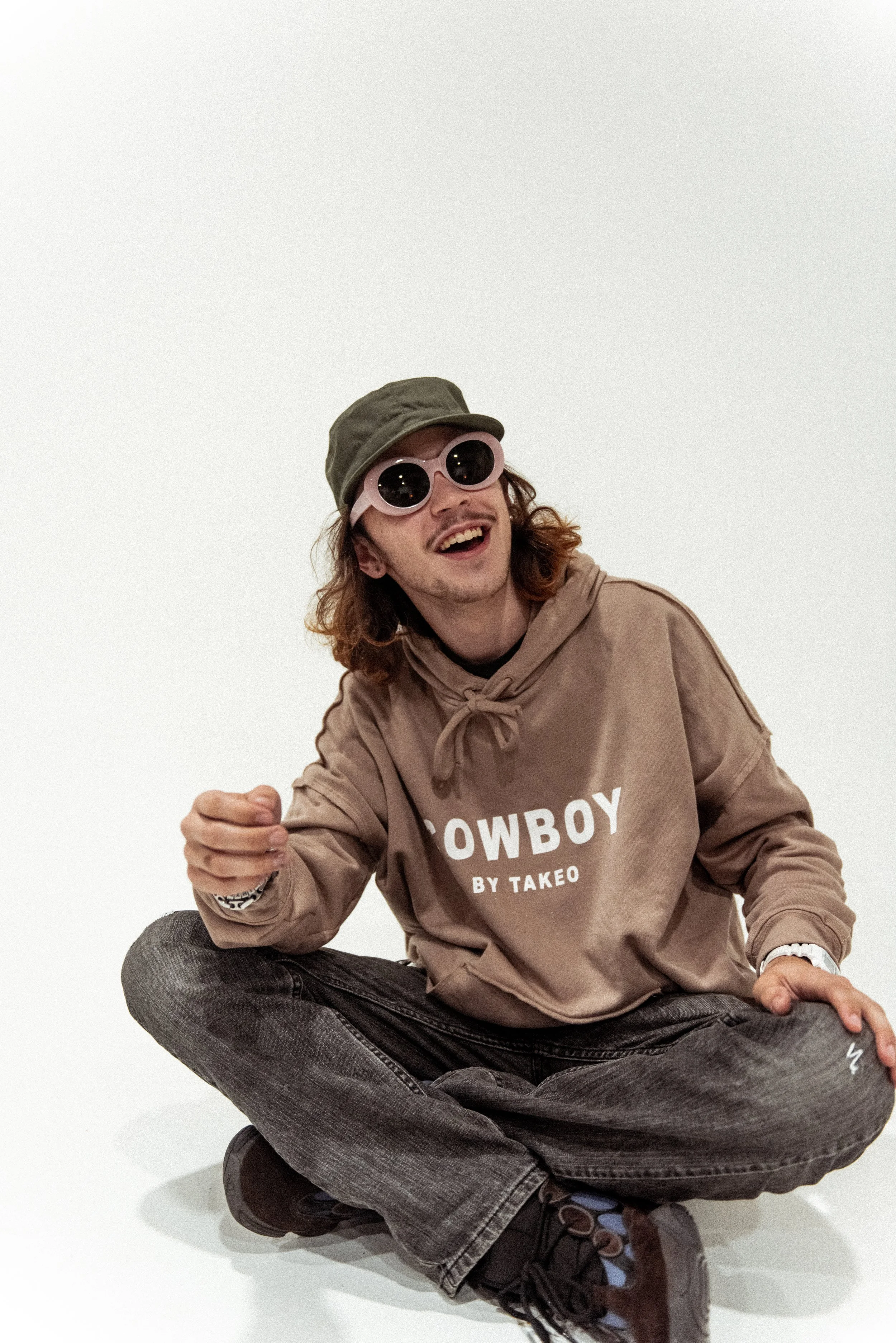 A young man with shoulder-length curly hair, wearing a beige hoodie with 'COWBOY BY TAKEO' printed on it, black jeans, brown shoes, pink sunglasses, and a gray cap, sitting on the floor against a white background, smiling and laughing.