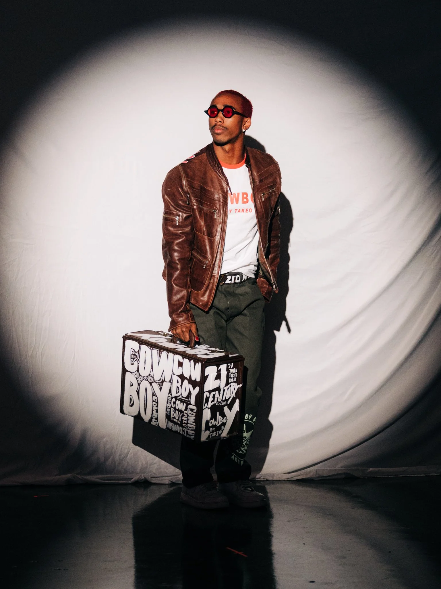 A man with reddish hair and sunglasses holding a large black and white shopping bag standing in a spotlight against a plain backdrop.