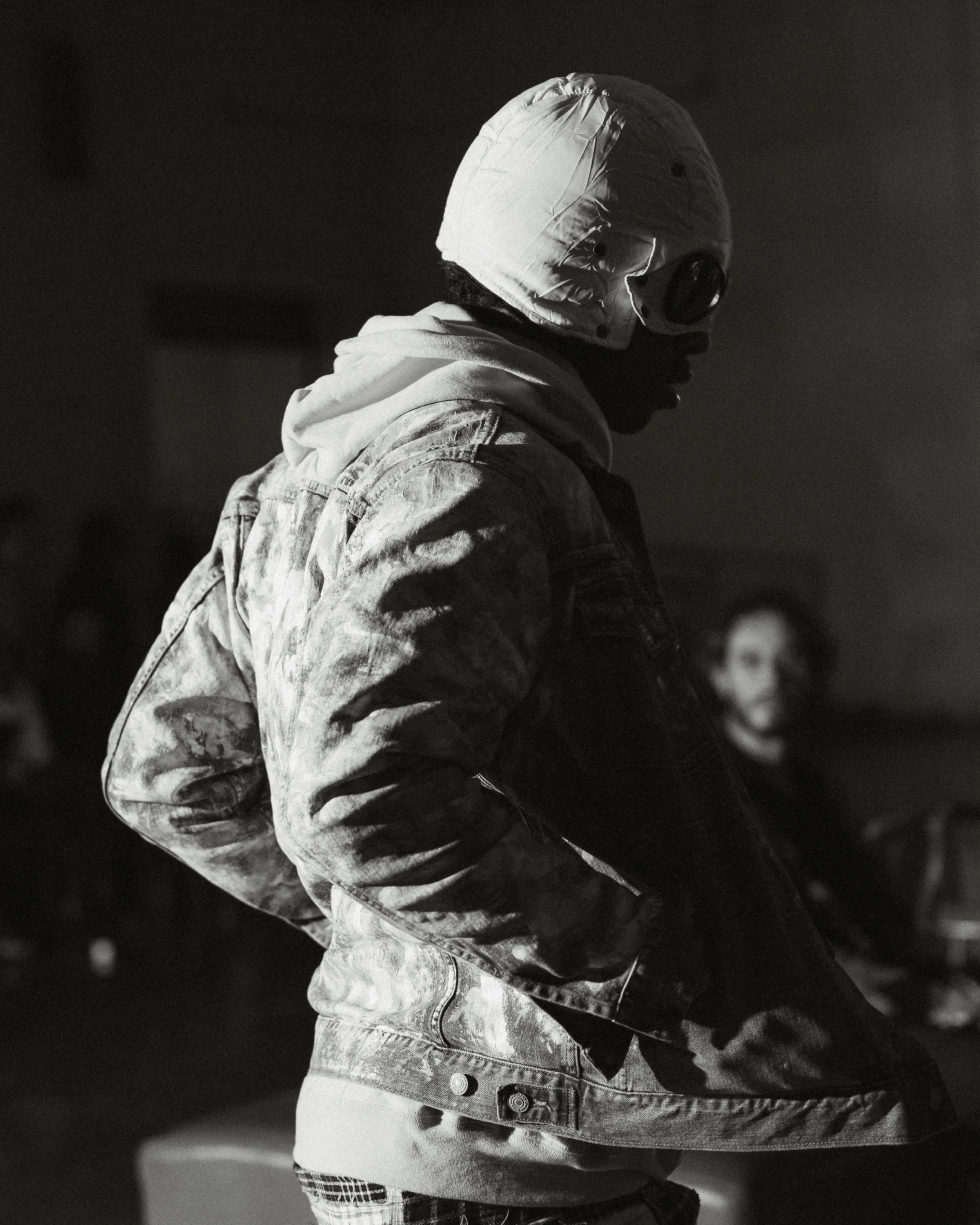 Person wearing a hoodie, denim jacket, and protective goggles, standing with hands in jacket pockets, in a dimly lit indoor space.