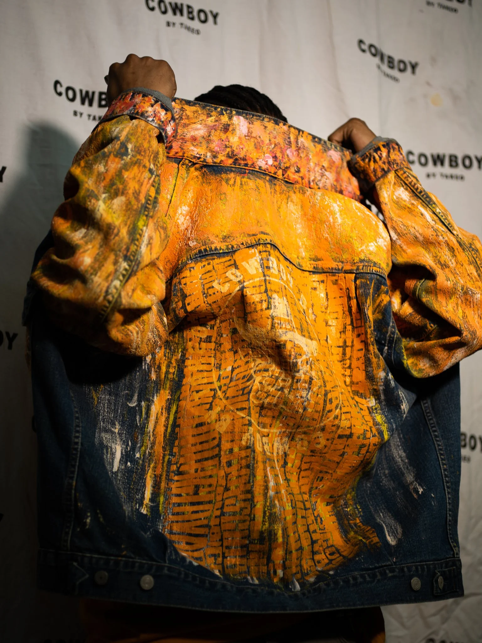 A person is wearing a vibrant, painted denim jacket with orange, black, and yellow abstract patterns and city map designs on the back, standing in front of a backdrop with the words "Cowboy by Takeo" repeatedly printed.