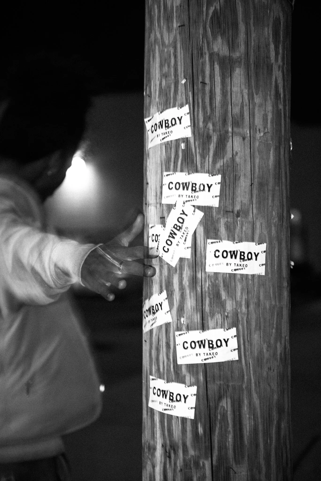 A person pasting multiple stickers that say 'COWBOY' on a wooden utility pole at night.