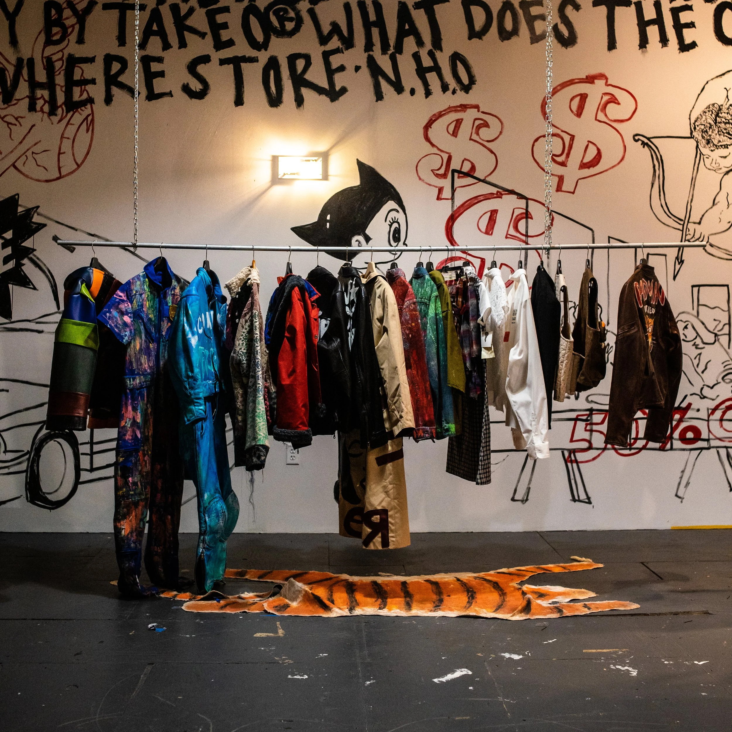 Clothing rack with colorful jackets and shirts inside a store, with graffiti-style wall art including dollar signs, a cartoon girl, and other drawings in the background.