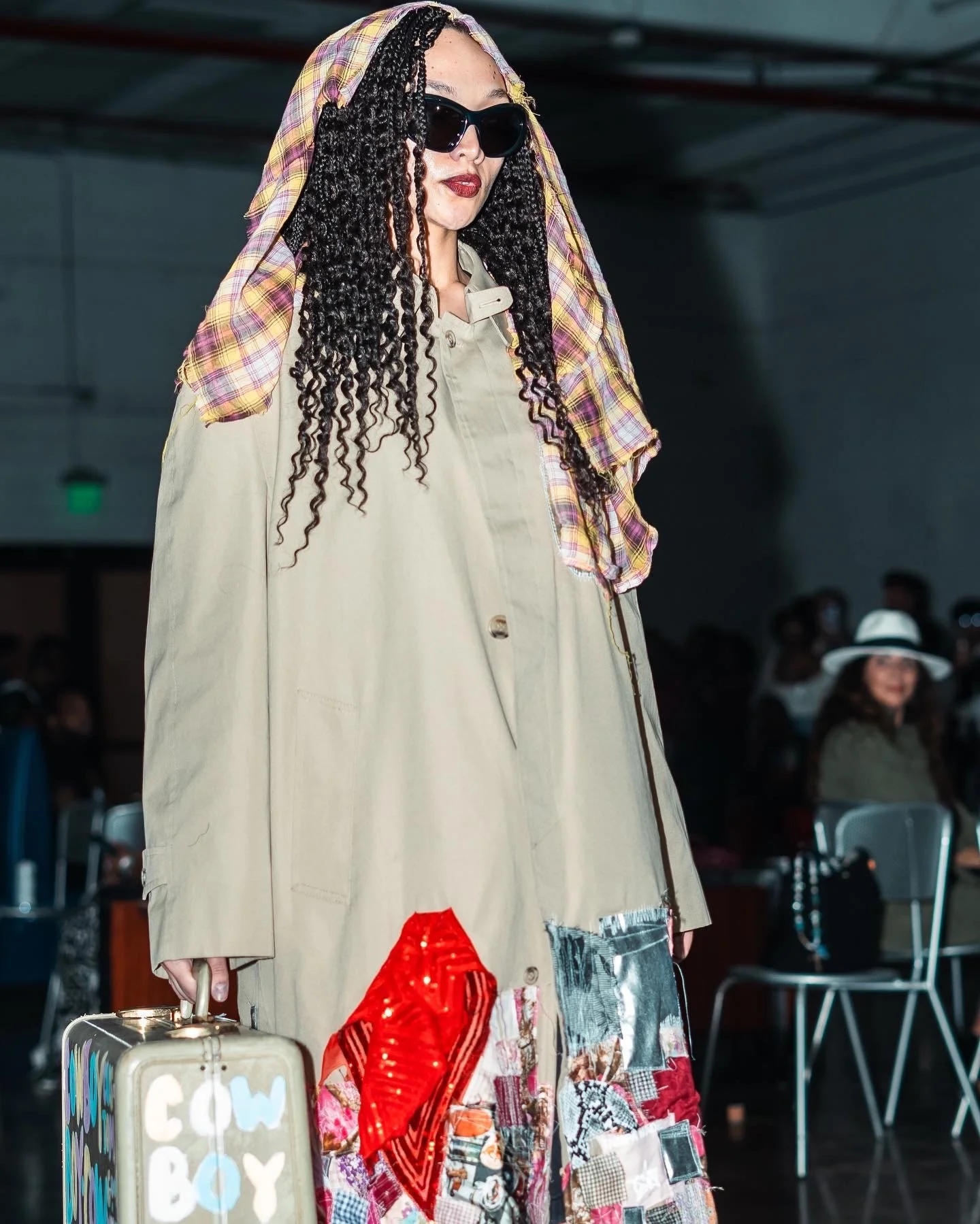 A woman with long, curly black hair, wearing black sunglasses, a beige trench coat with a patchwork skirt, and holding a silver suitcase with the words 'COWBOY' written on it. She has a plaid cloth draped over her head.