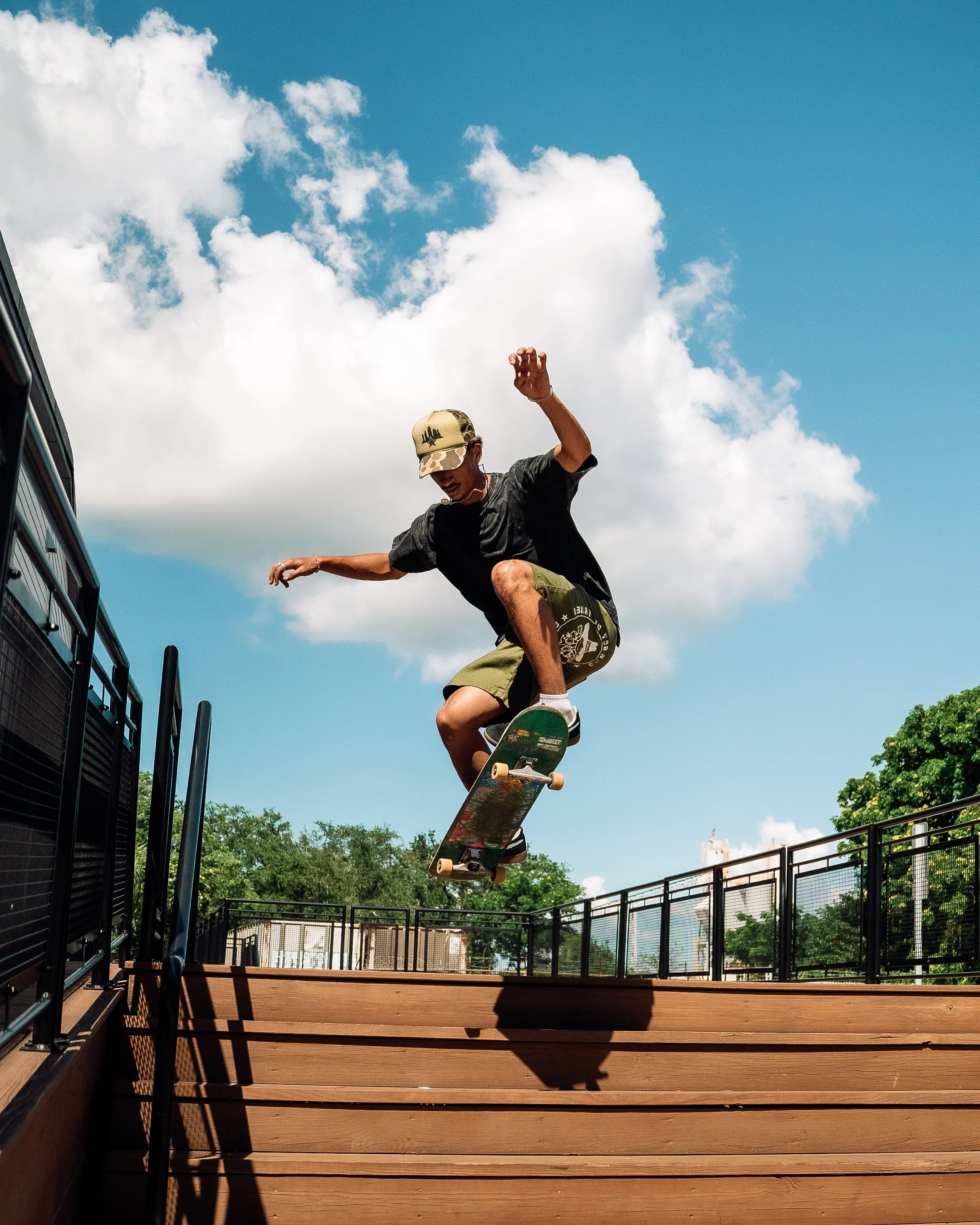 Skateboarder wearing a beige hat, black T-shirt, and green shorts performing an ollie off wooden stairs at an outdoor skate park on a sunny day with a blue sky and white clouds.