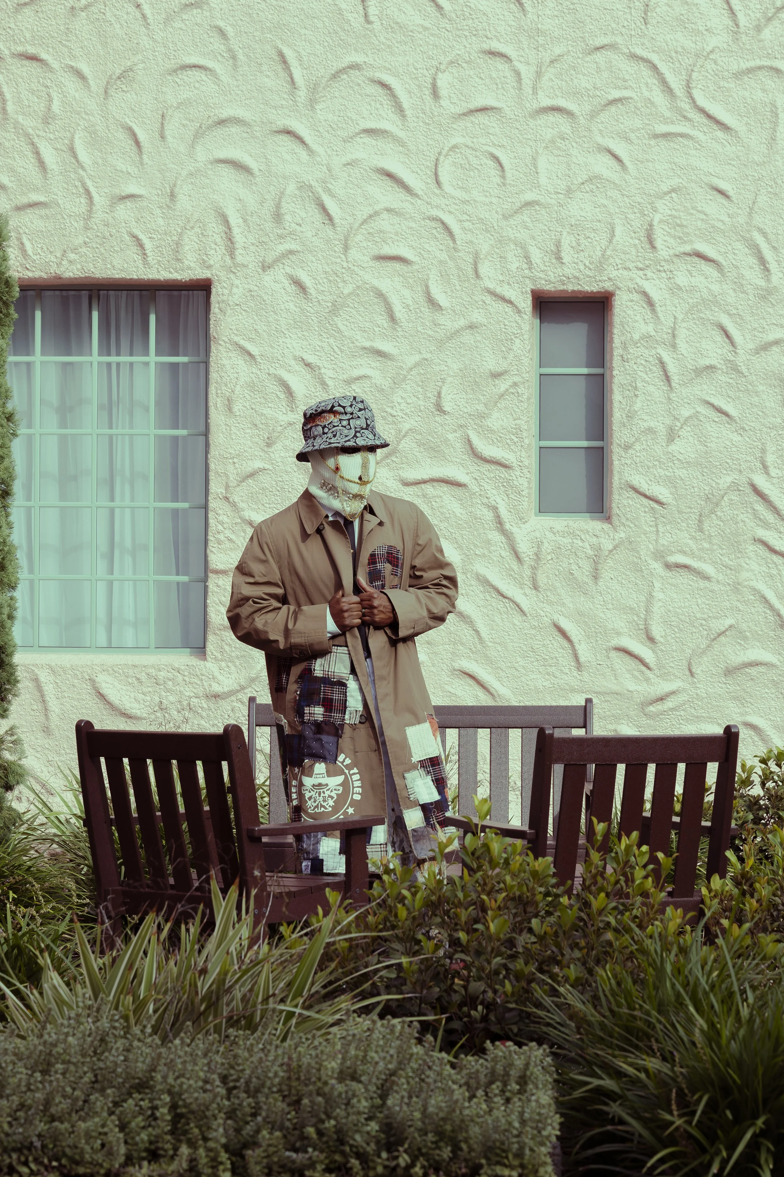 A mannequin wearing a beige coat, checkered patchwork pants, and a colorful bucket hat, standing in front of a textured yellow wall with two small windows, surrounded by green plants and seated on two black garden benches.
