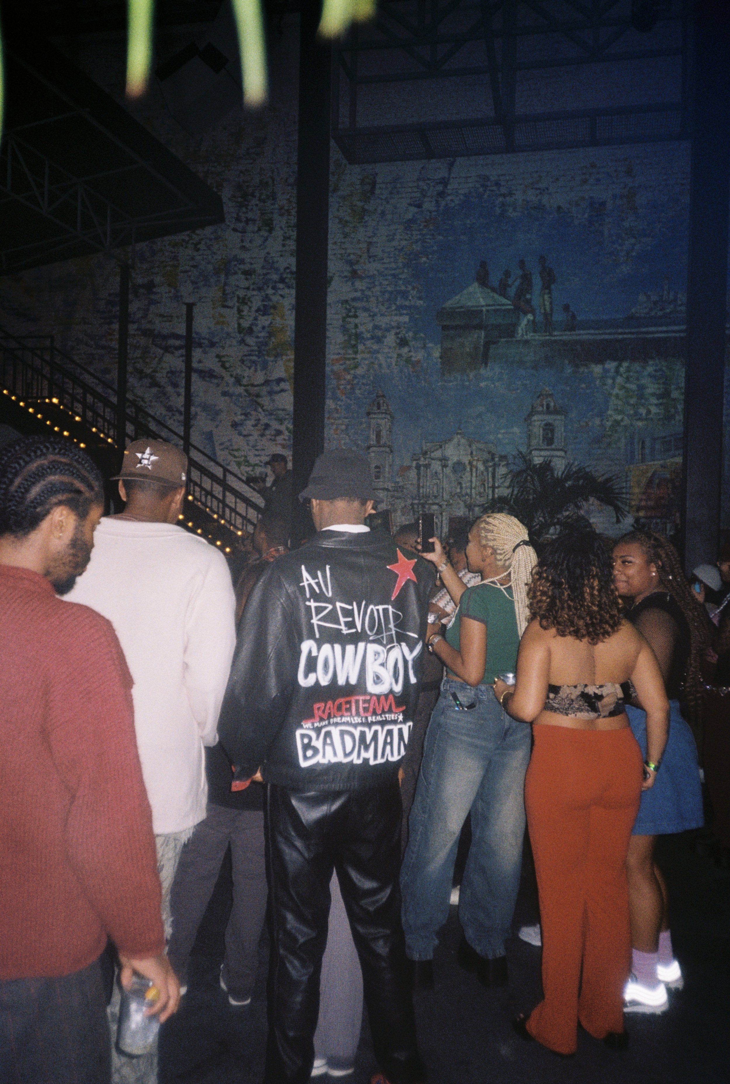 Group of people at a nightlife event, with a mural in the background and one person wearing a black leather jacket with handwritten-style text about rebellion and attitude.