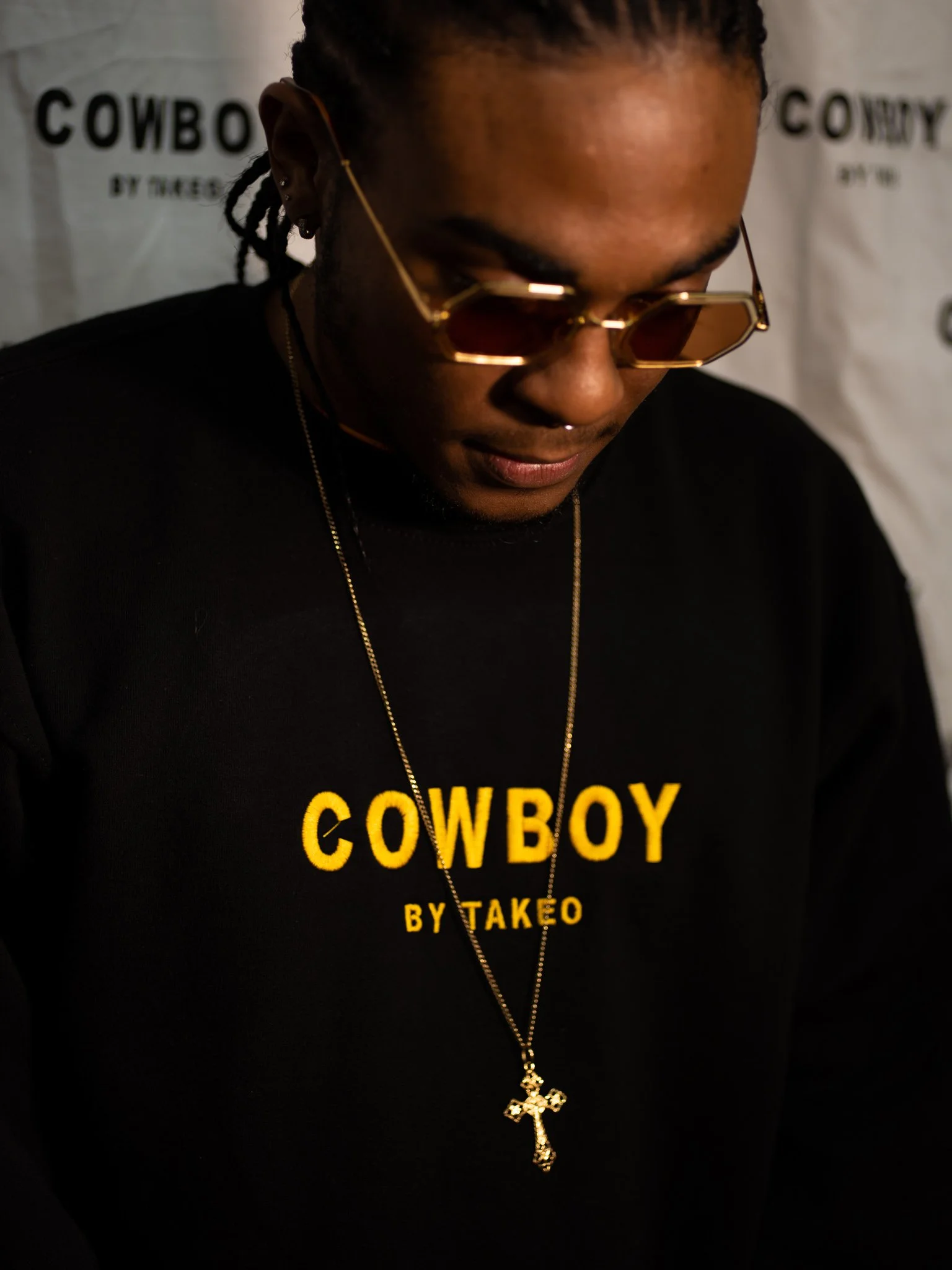 A man wearing gold sunglasses, a black sweatshirt with 'COWBOY BY TAKEO' in yellow lettering, a gold necklace with a cross pendant, and earrings. He is looking down in front of a white background with partial text.