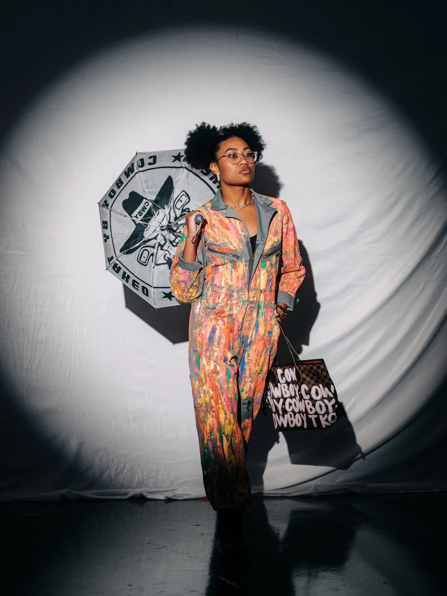 A woman with curly hair and glasses standing against a gray background, holding a patterned umbrella in one hand and a large shopping bag in the other, wearing a colorful paint-splattered jumpsuit.