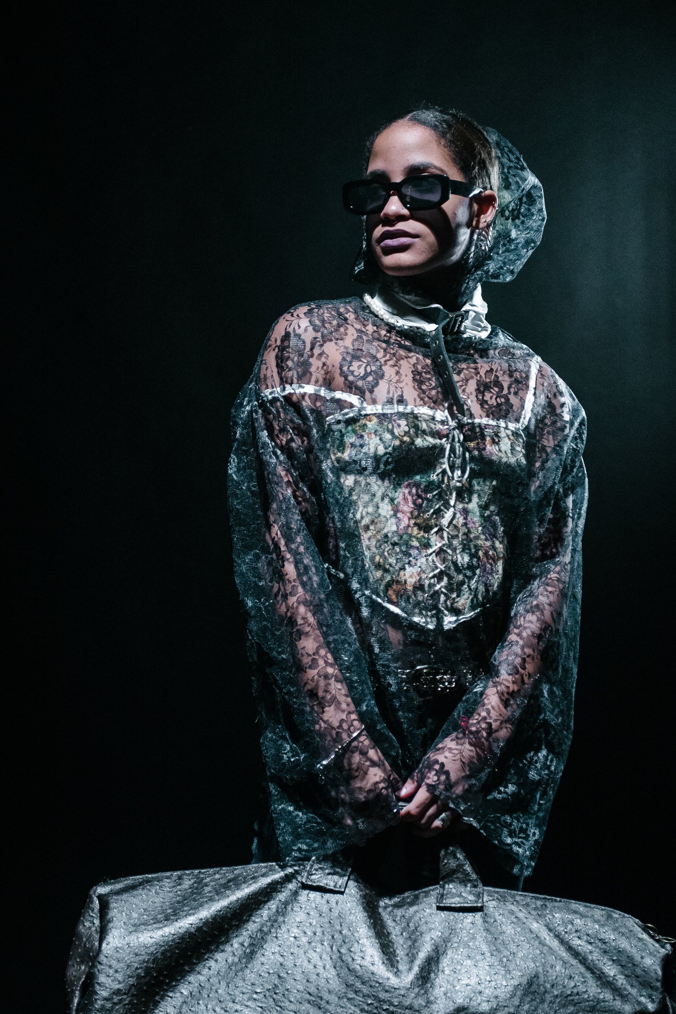 A woman wearing sunglasses, lace top, headscarf, standing with a large bag.