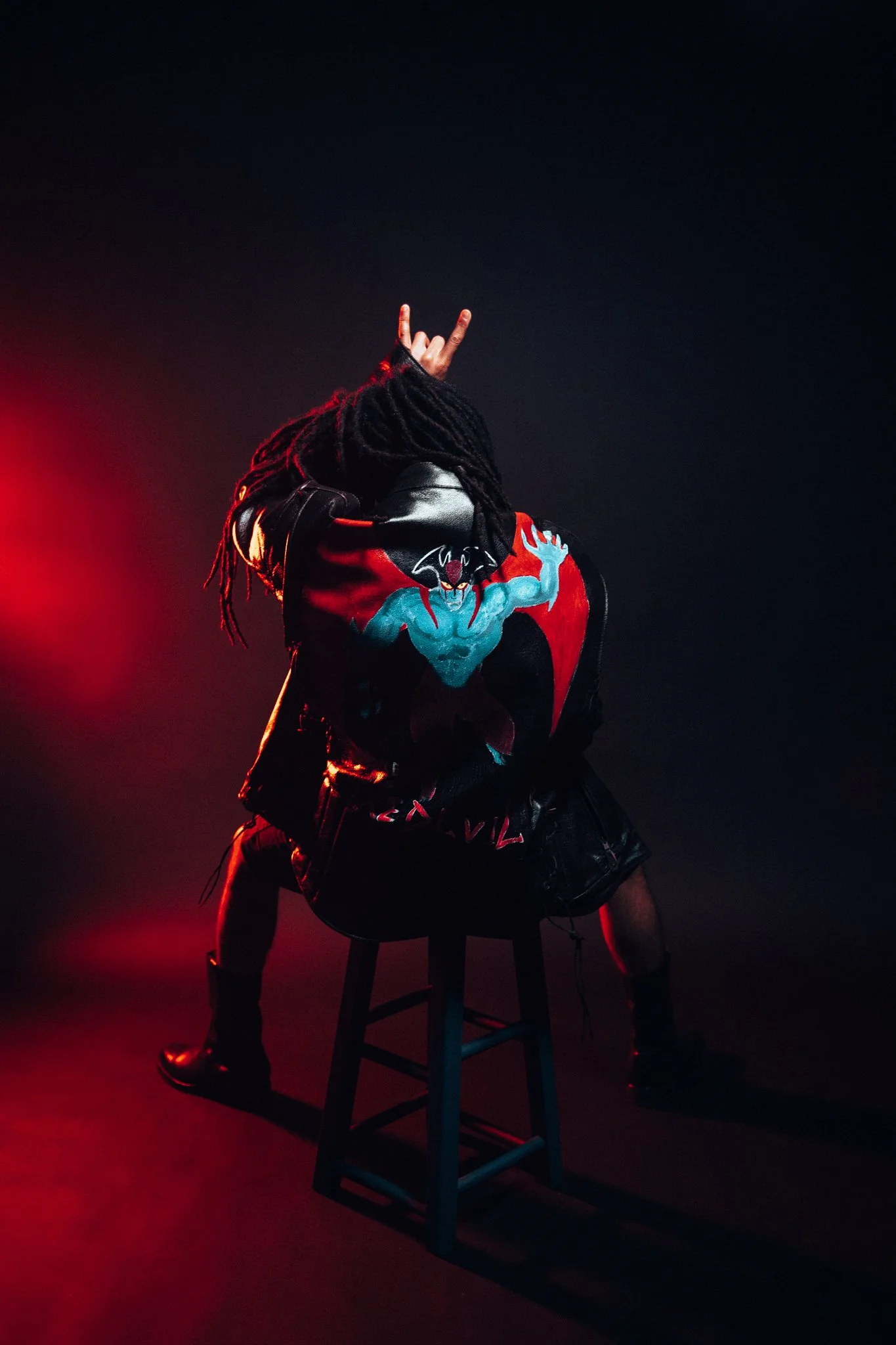 Person with dreadlocks sitting on a stool, wearing a colorful jacket with a graphic of a creature with wings and a bat-like face, posing with one hand making rock and roll gestures in a dark setting with red lighting.