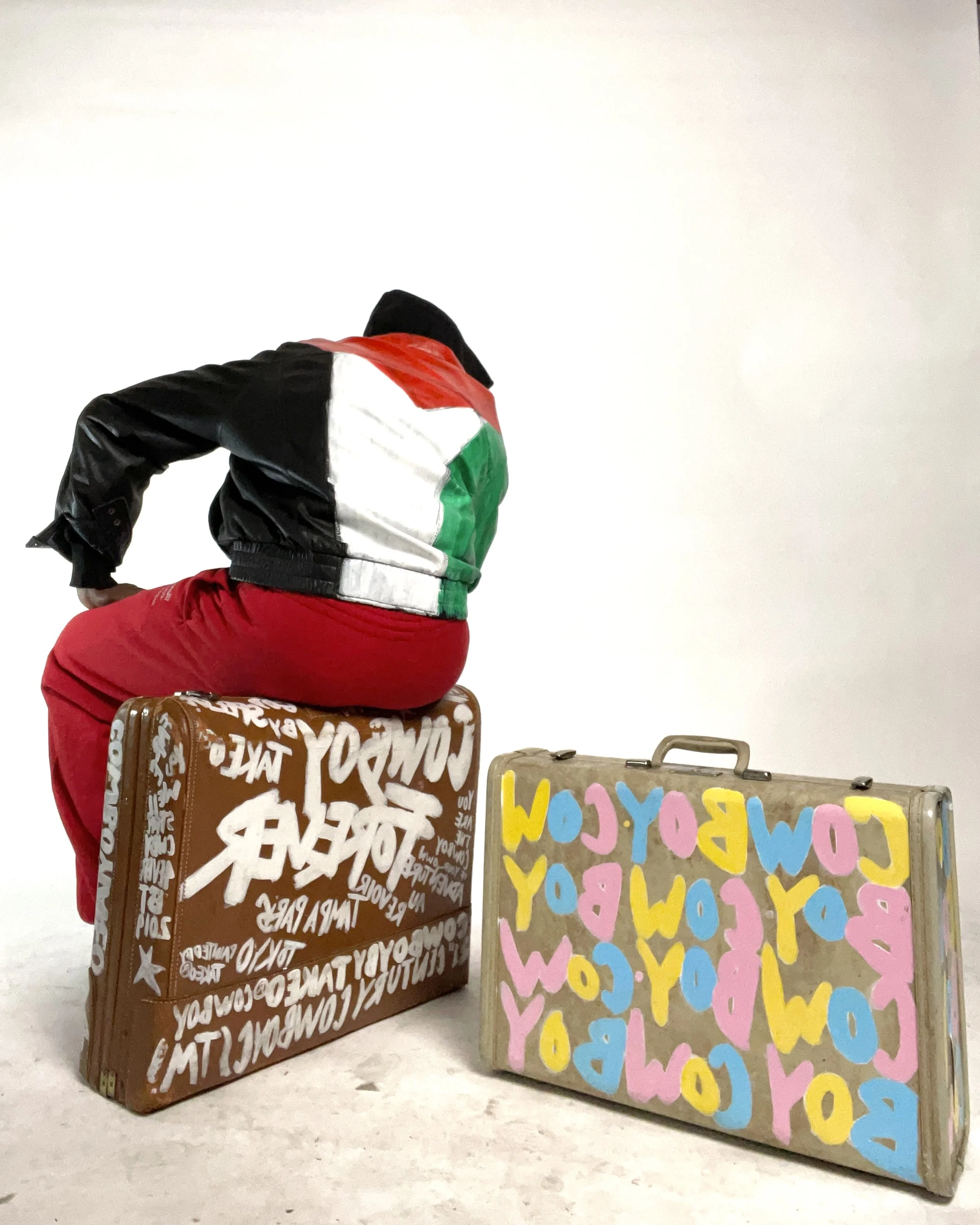 A person with their back turned, sitting on a decorated suitcase, wearing a jacket with the colors of the United Arab Emirates flag, and surrounded by colorful, graffiti-style suitcases.