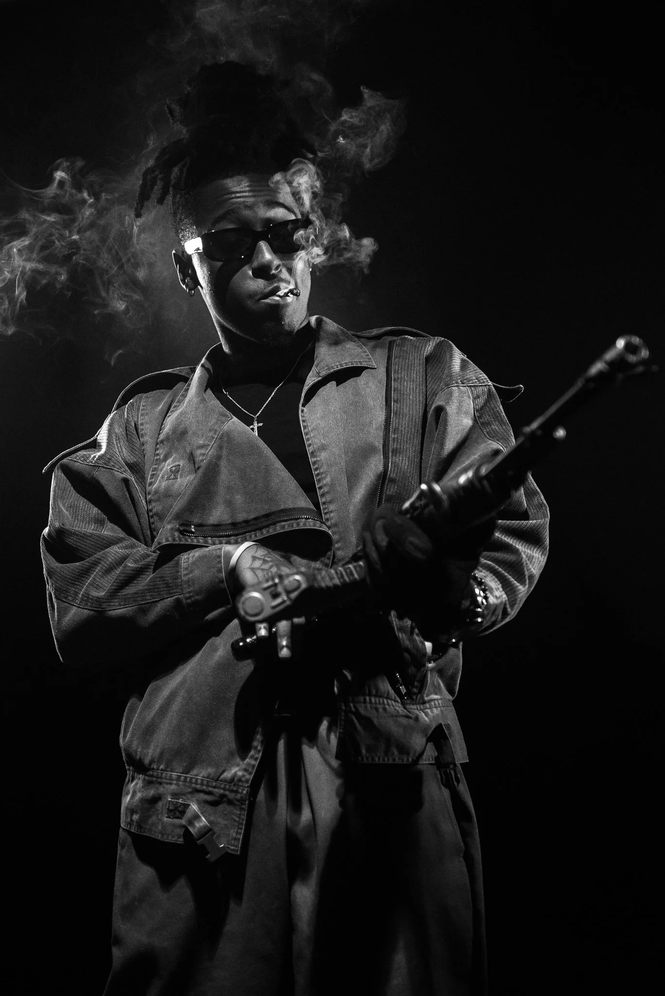 A black and white photo of a man with dreadlocks and sunglasses holding a gun, wearing a jacket and a cross necklace, with smoke around his head.